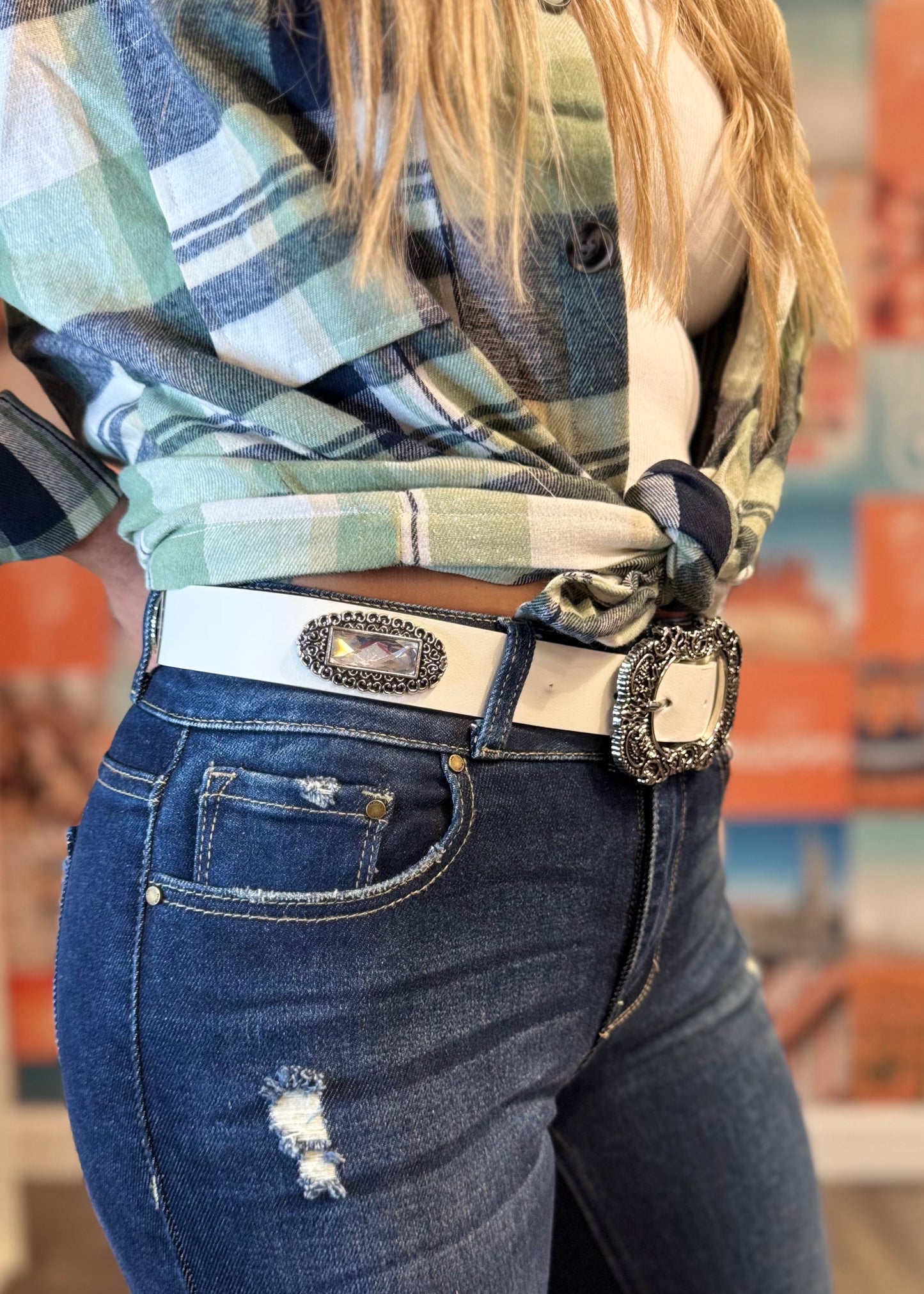 A close-up image of a woman wearing casual, trendy attire. She is dressed in a tied, green and blue plaid flannel shirt over a white ribbed tank top. Her outfit is paired with dark blue distressed skinny jeans featuring small rips and a white belt with a decorative silver buckle.