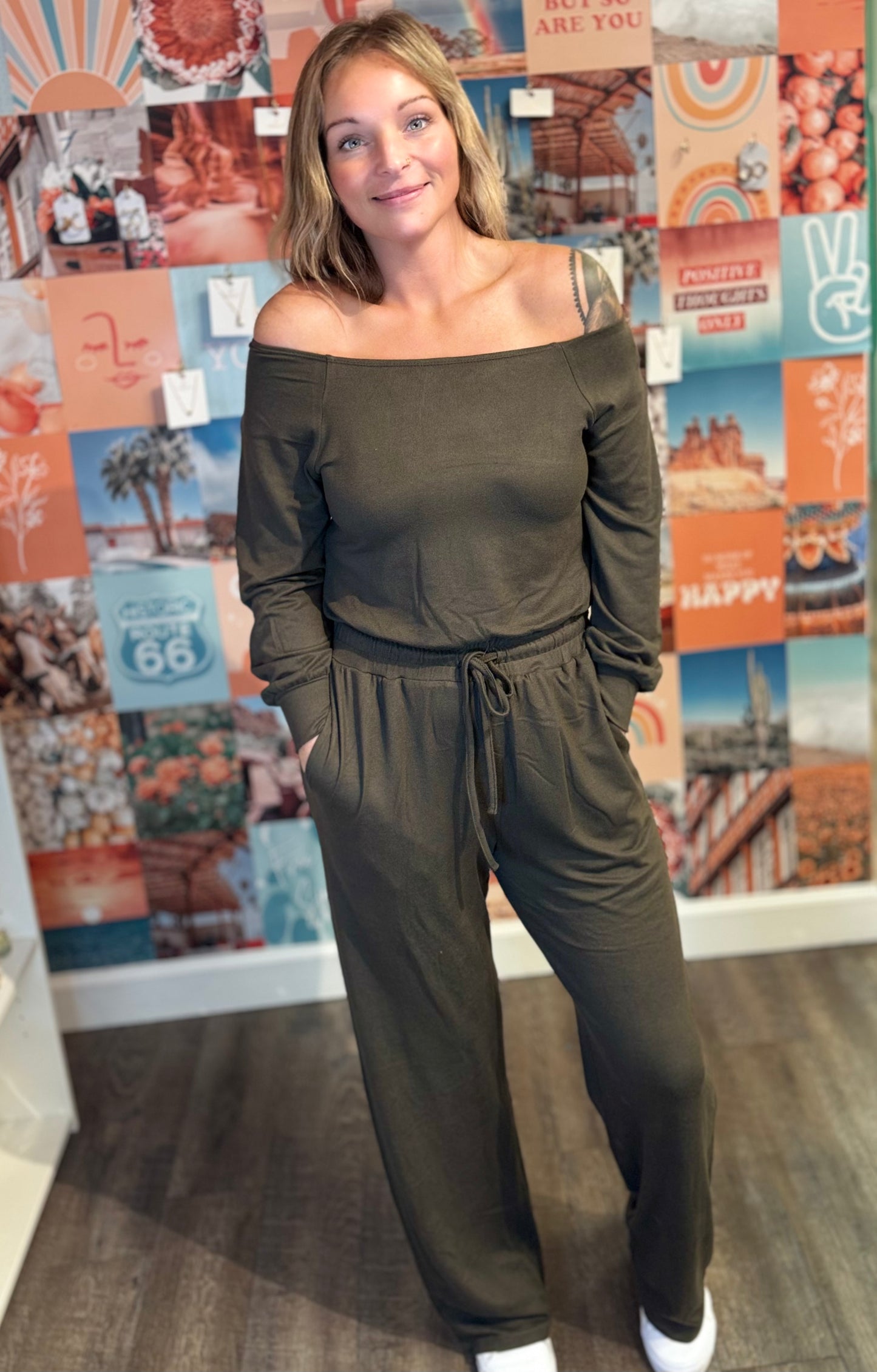 A woman wearing an olive-green off-the-shoulder jumpsuit featuring long sleeves, an elastic waistband with a drawstring, and wide-leg pants. The outfit includes side pockets, offering a casual yet chic look. She is standing indoors on a wooden floor, with a vibrant collage wall in the background featuring various colorful prints, patterns, and inspirational imagery. She pairs the outfit with white sneakers for a relaxed and comfortable style.