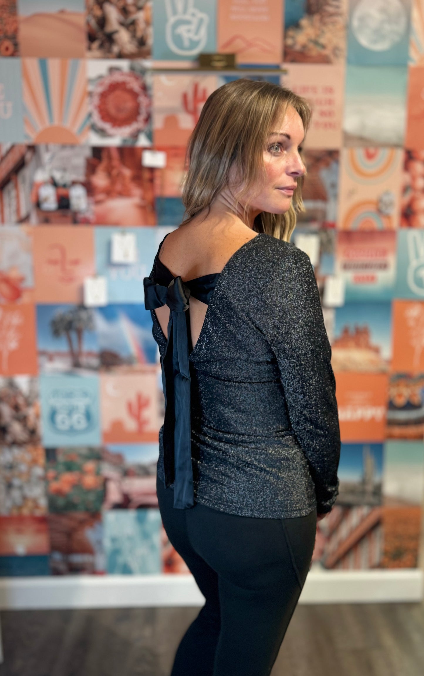Elegant black shimmer top featuring a low back design adorned with a large satin bow detail. The long bow ties add a touch of sophistication, perfect for evening wear or special occasions. The glittery fabric enhances its chic appeal, complementing the fitted silhouette.