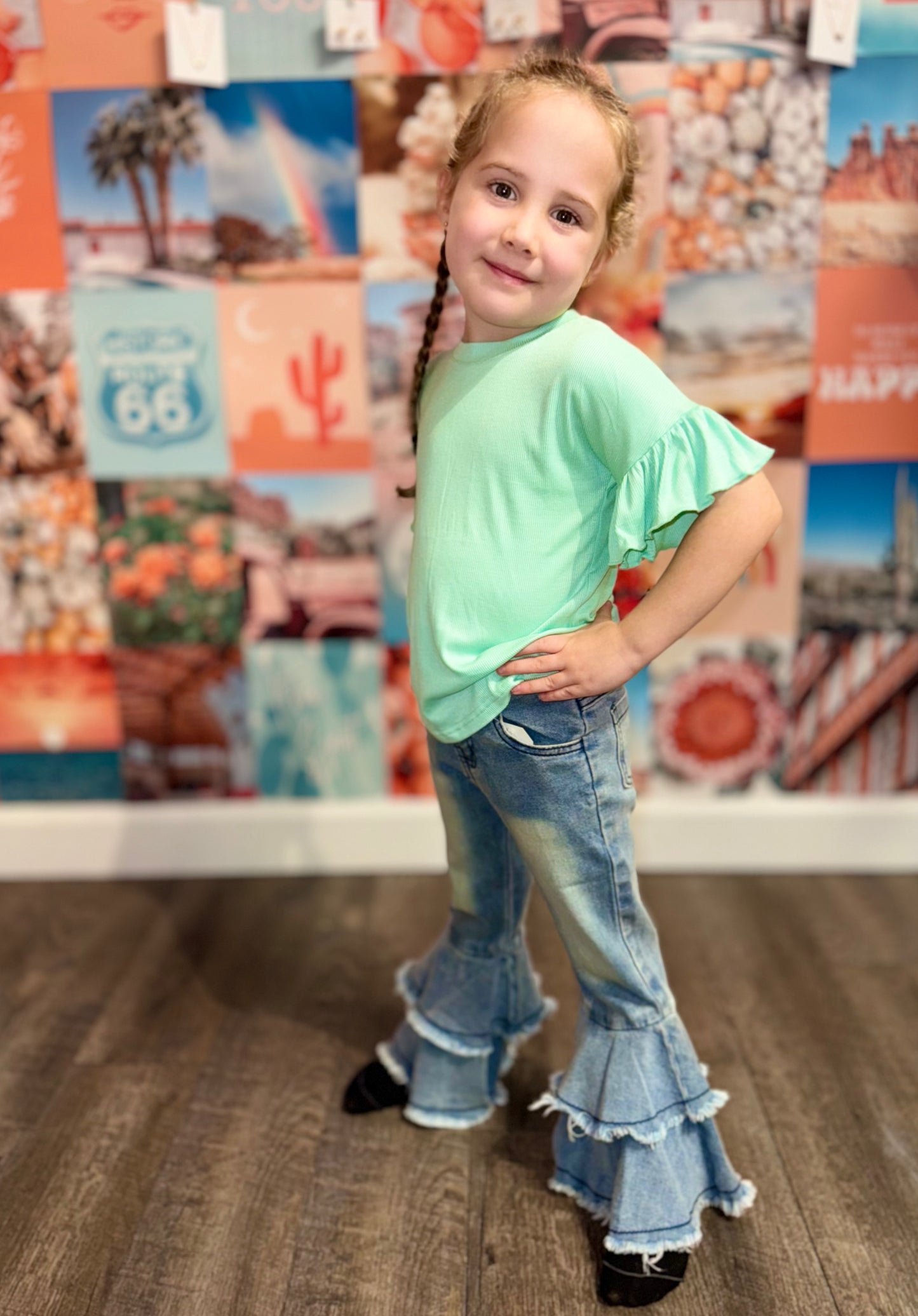 Little girl posing confidently in front of a colorful boho wall collage wearing jean bell-bottoms and a teal aqua top with ruffled sleeves. A trendy, playful outfit perfect for stylish kids who love fun and comfortable fashion