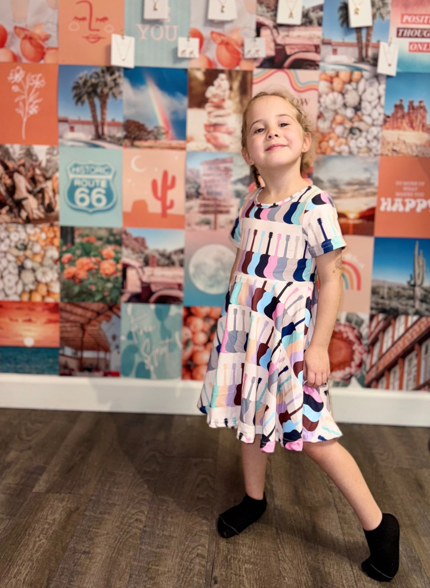 Little girl wearing a colorful Taylor Swift-inspired dress with guitar patterns, standing confidently in front of a vibrant boho wall collage featuring peach, teal, and pink tones. Perfect twirl dress for kids who love music and fun fashion.
