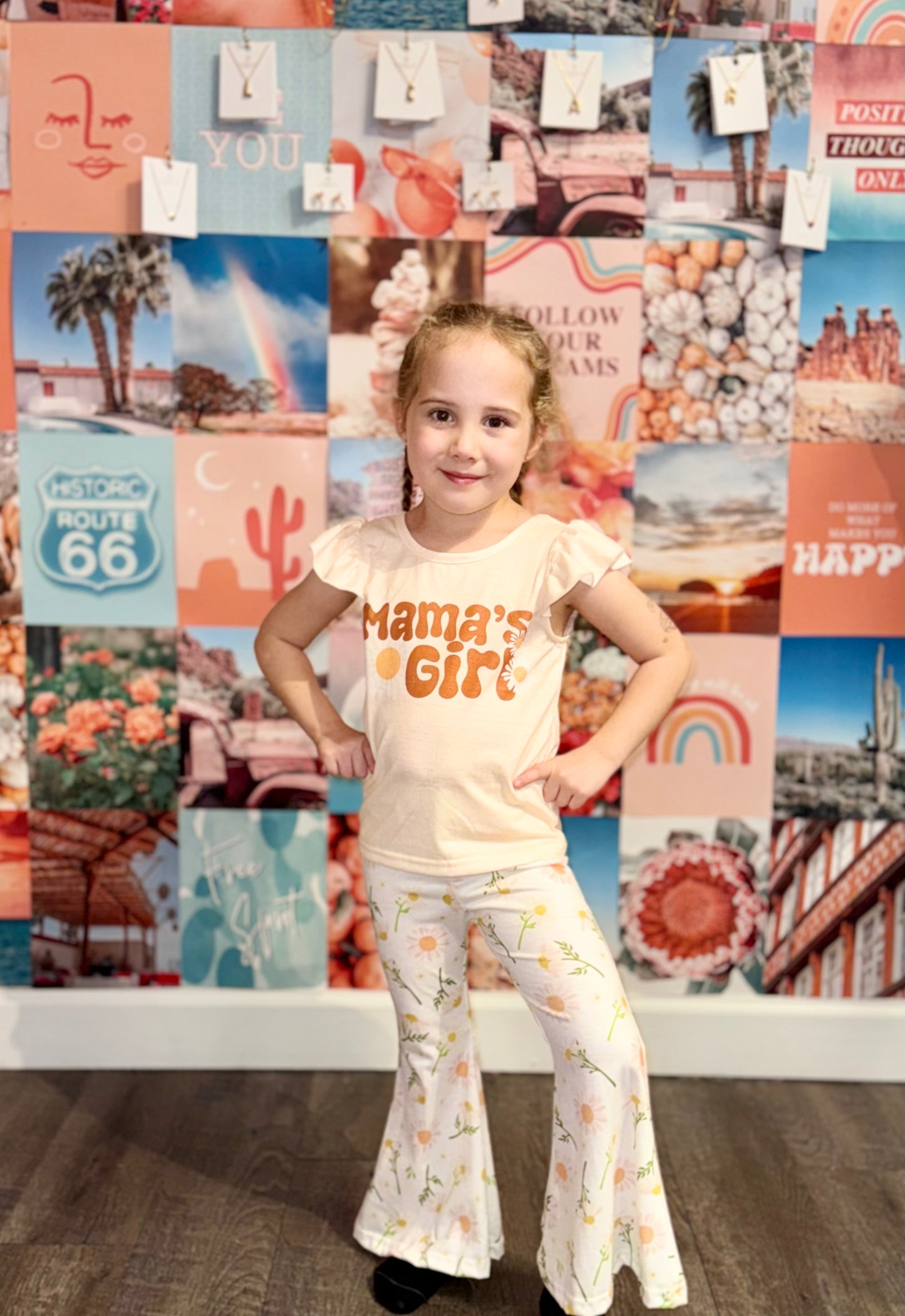 Little girl posing in a peach ‘Mama’s Girl’ shirt and white floral bell-bottom pants, standing confidently in front of a colorful boho wall collage with teal, peach, and pink tones. Perfect stylish outfit for kids who love playful yet trendy fashion