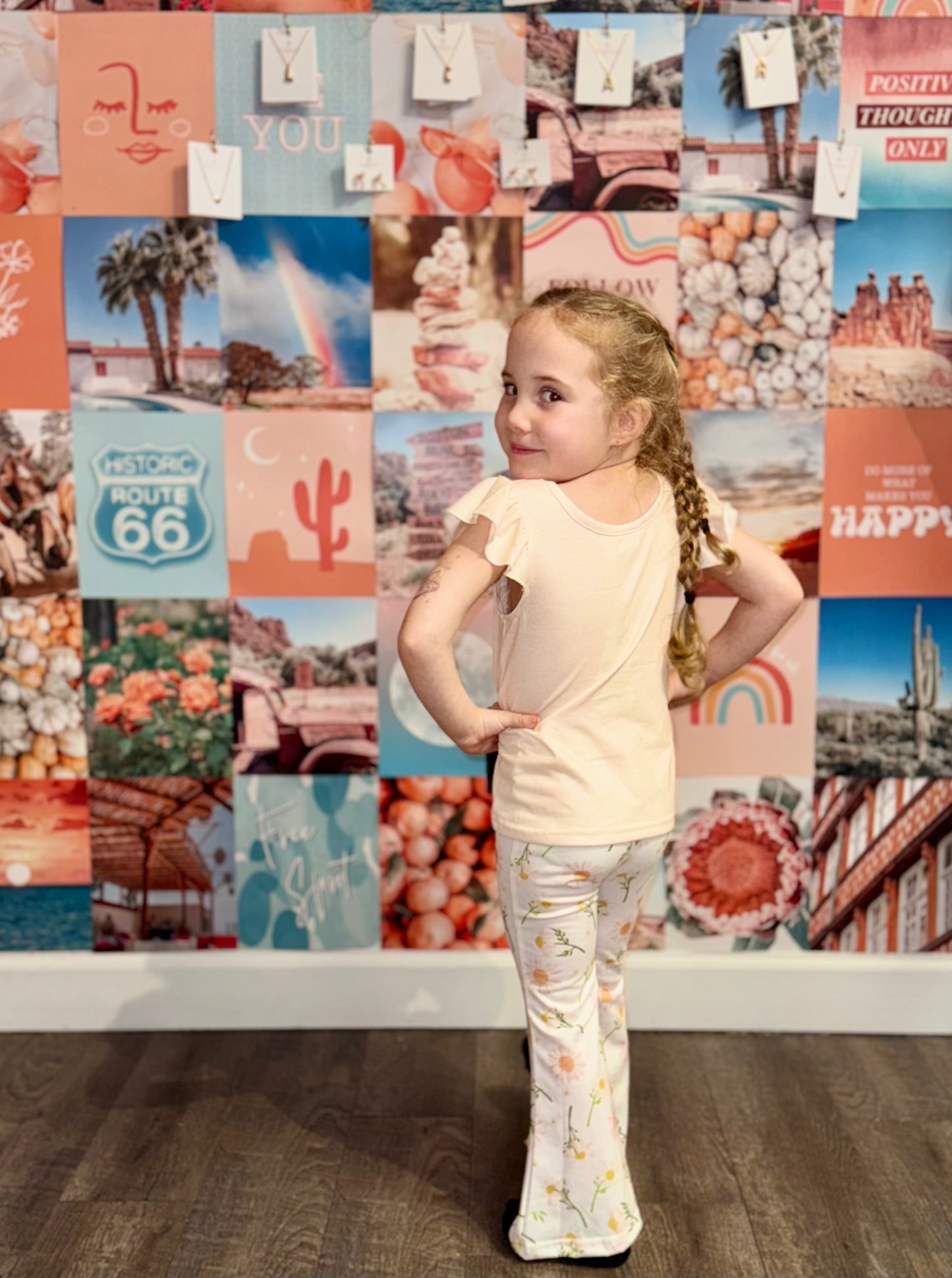 Little girl posing in a peach ‘Mama’s Girl’ shirt and white floral bell-bottom pants, standing confidently in front of a colorful boho wall collage with teal, peach, and pink tones. Perfect stylish outfit for kids who love playful yet trendy fashion
