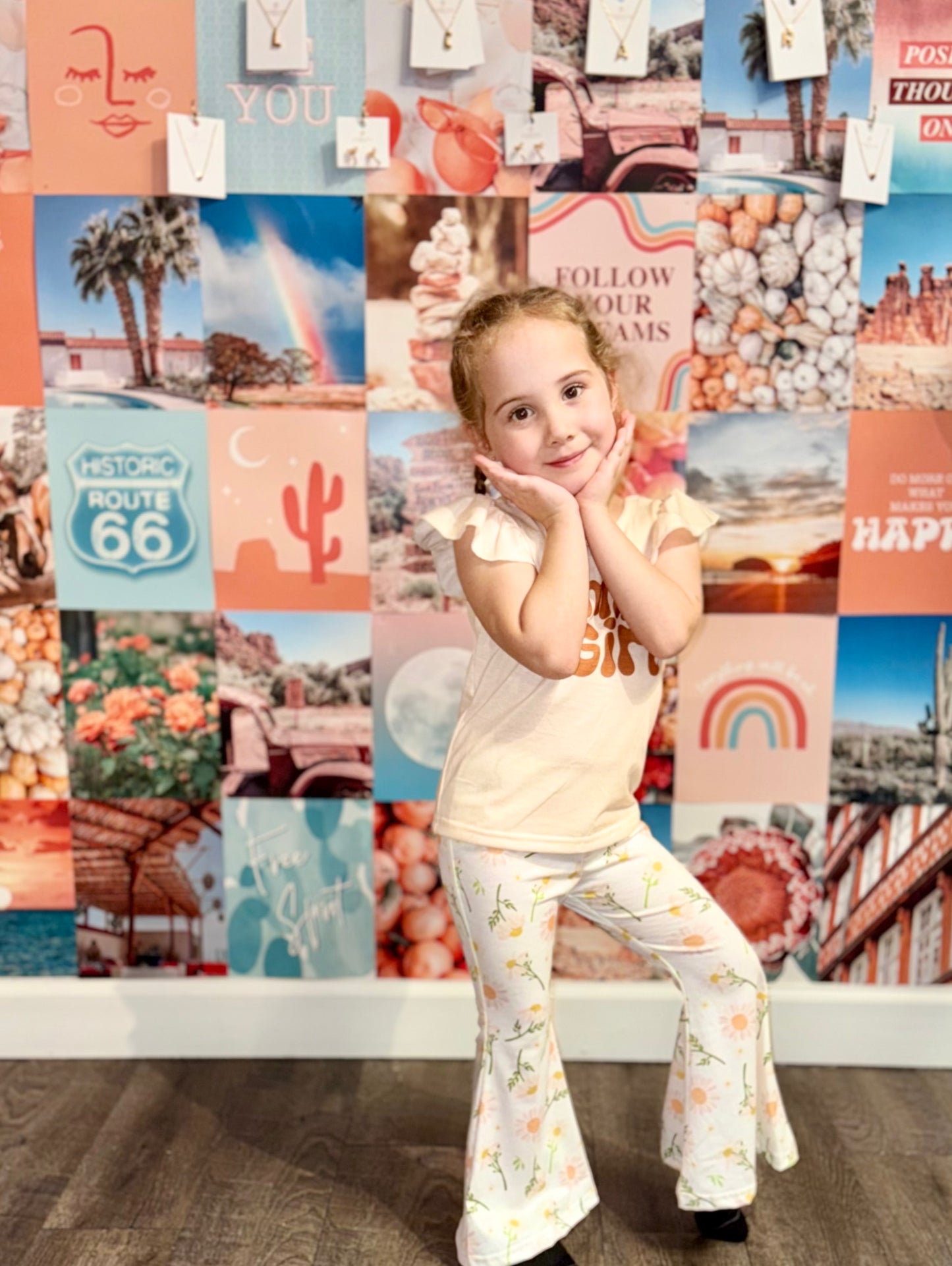 Little girl posing in a peach ‘Mama’s Girl’ shirt and white floral bell-bottom pants, standing confidently in front of a colorful boho wall collage with teal, peach, and pink tones. Perfect stylish outfit for kids who love playful yet trendy fashion