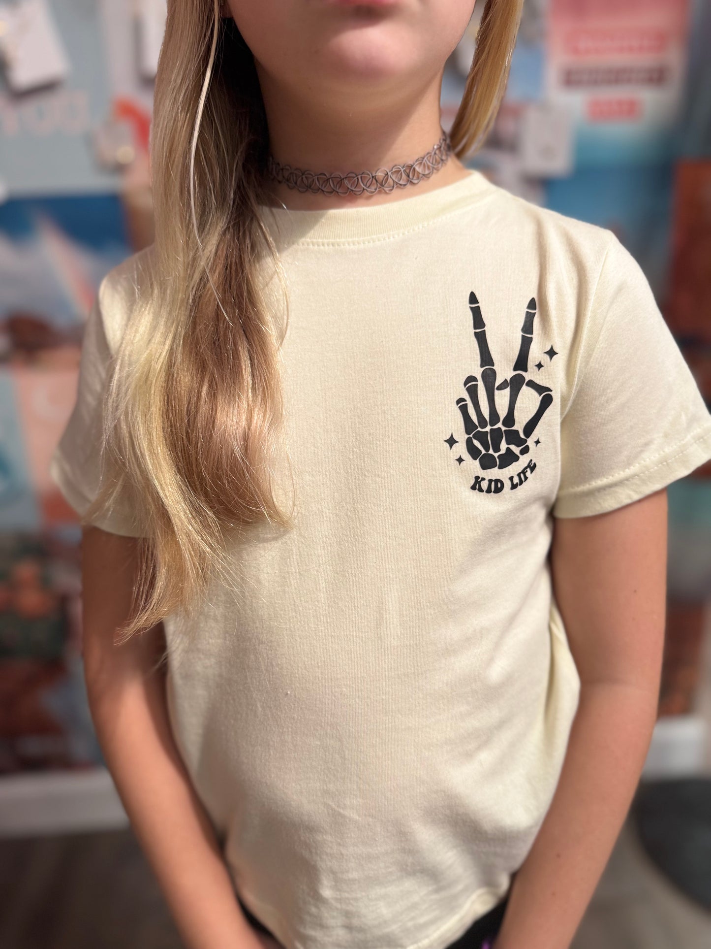 Youth cream-colored t-shirt with a black skeleton hand design forming a peace sign, accompanied by the text 'Kid Life.' Styled with a trendy choker necklace and worn by a young girl, showcasing a fun and edgy vibe perfect for casual outfits.