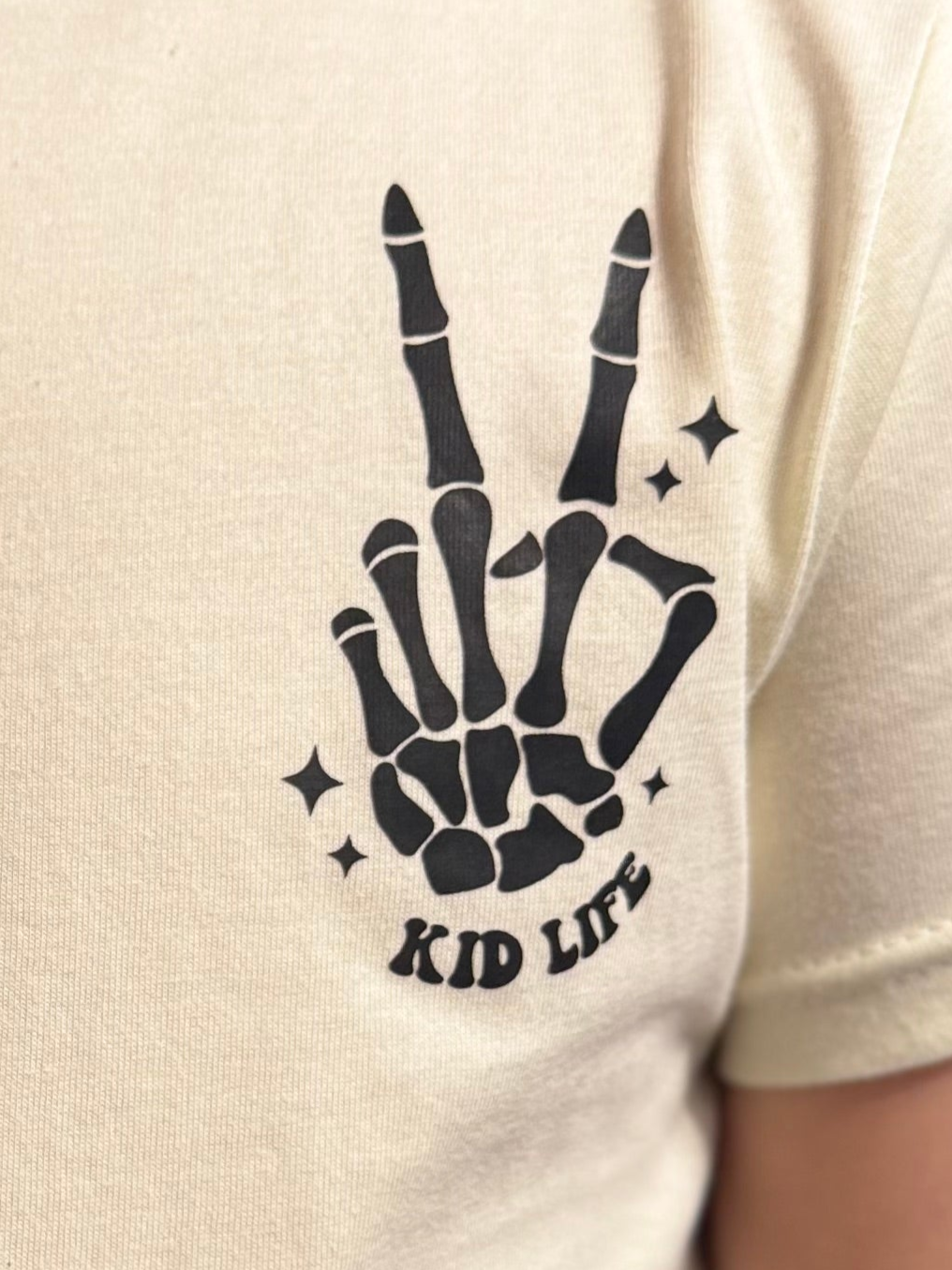 Youth cream-colored t-shirt with a black skeleton hand design forming a peace sign, accompanied by the text 'Kid Life.' Styled with a trendy choker necklace and worn by a young girl, showcasing a fun and edgy vibe perfect for casual outfits.