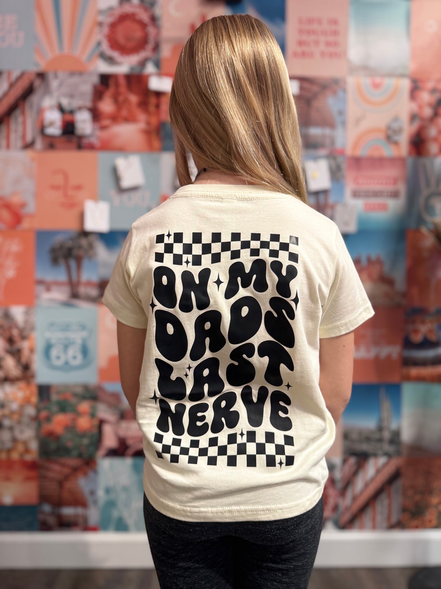 Cream-colored youth t-shirt featuring bold black text that reads 'On My Dad's Last Nerve' in a playful retro font, with checkerboard accents and star details. The design is showcased on a young girl standing against a colorful photo collage wall, emphasizing the shirt’s humorous and trendy appeal.