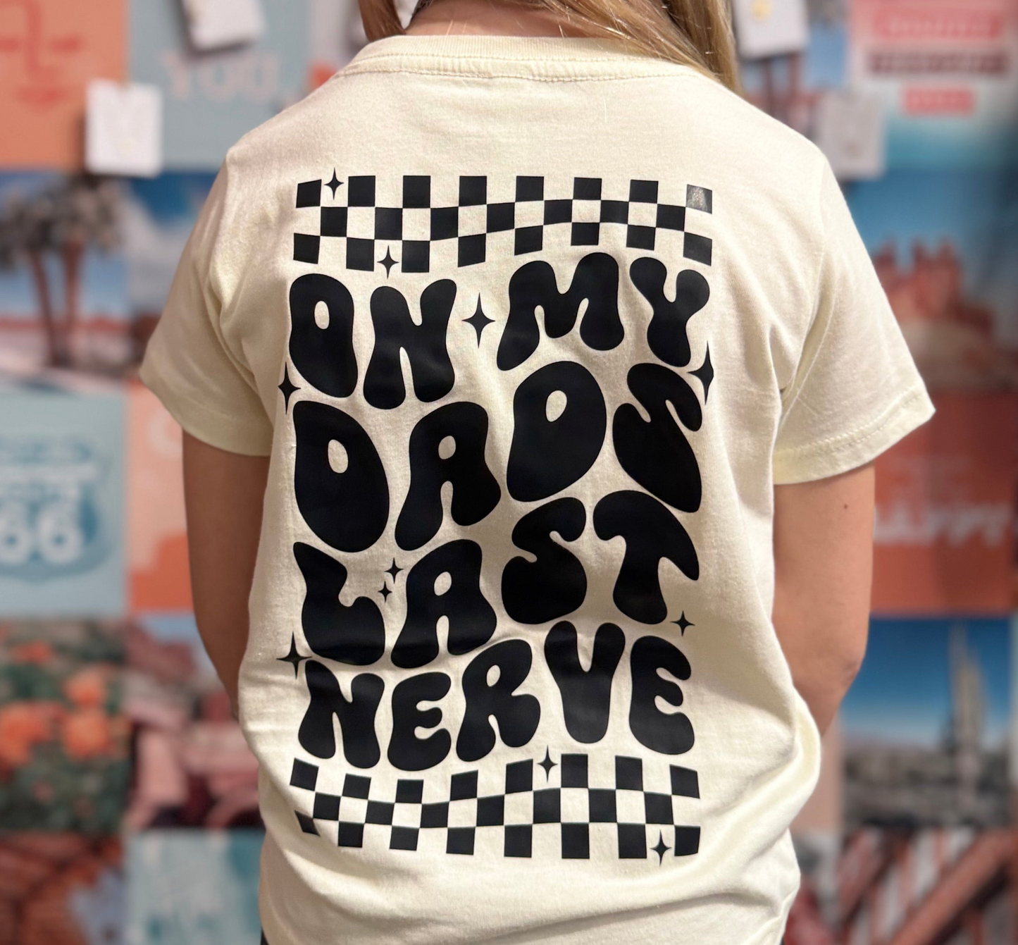Cream-colored youth t-shirt featuring bold black text that reads 'On My Dad's Last Nerve' in a playful retro font, with checkerboard accents and star details. The design is showcased on a young girl standing against a colorful photo collage wall, emphasizing the shirt’s humorous and trendy appeal.