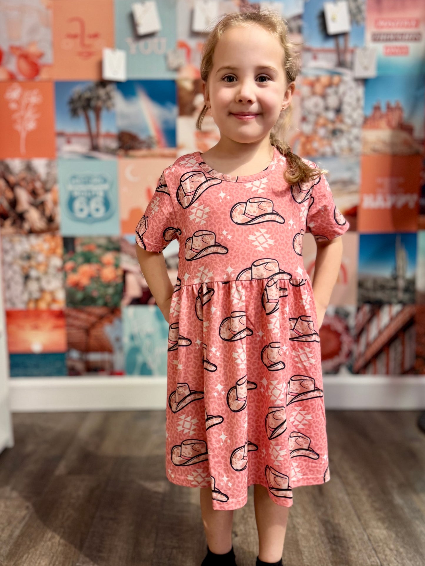 Young girl wearing a pink dress with a playful Western-inspired design featuring cowboy hats and geometric patterns. The dress has short sleeves and a flowy fit, styled against a colorful photo collage wall for a fun and casual look.