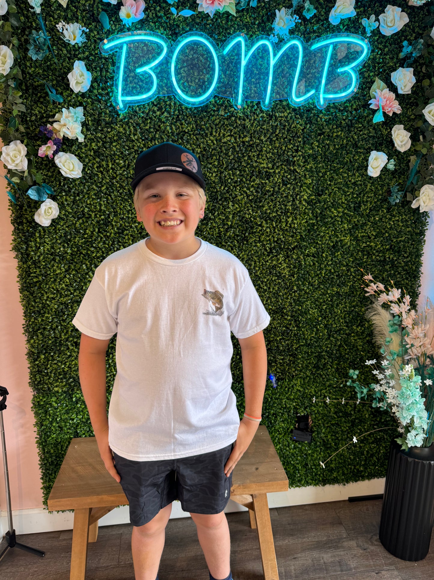Smiling young boy wearing a white T-shirt with a small bass fish graphic on the chest and black camouflage shorts. He is also wearing a black baseball cap with a tan deer skull patch. The backdrop features a vibrant green faux foliage wall adorned with white and pastel flowers and a glowing blue neon 'BOMB' sign, adding a fresh and lively vibe.