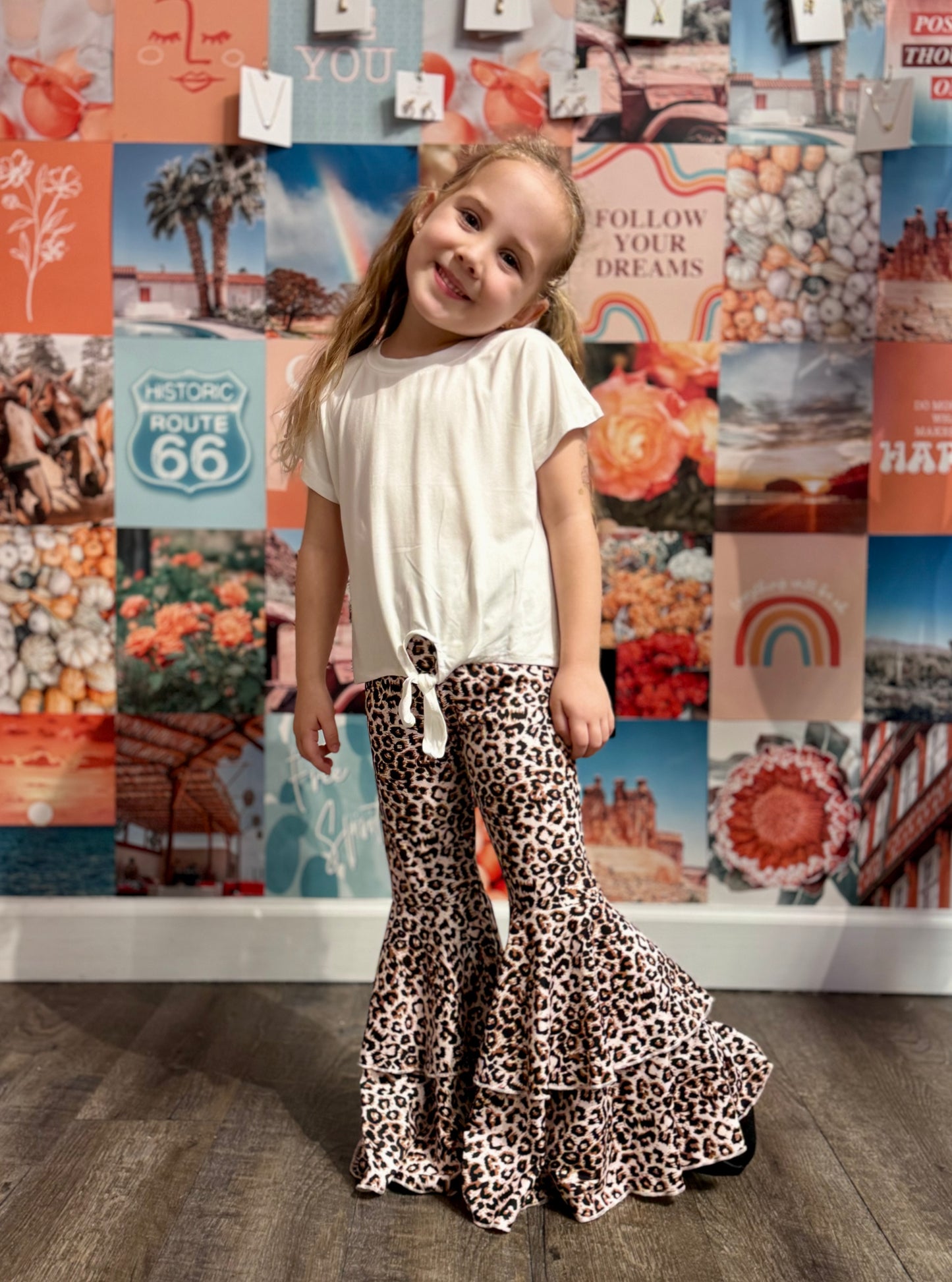 Smiling young girl posing confidently in a white tie-front T-shirt paired with flared bell-bottom pants featuring a bold leopard print pattern. The vibrant background is a colorful collage of retro and bohemian-inspired imagery, including Route 66 signs, rainbows, pumpkins, and desert landscapes, creating a playful and stylish aesthetic.