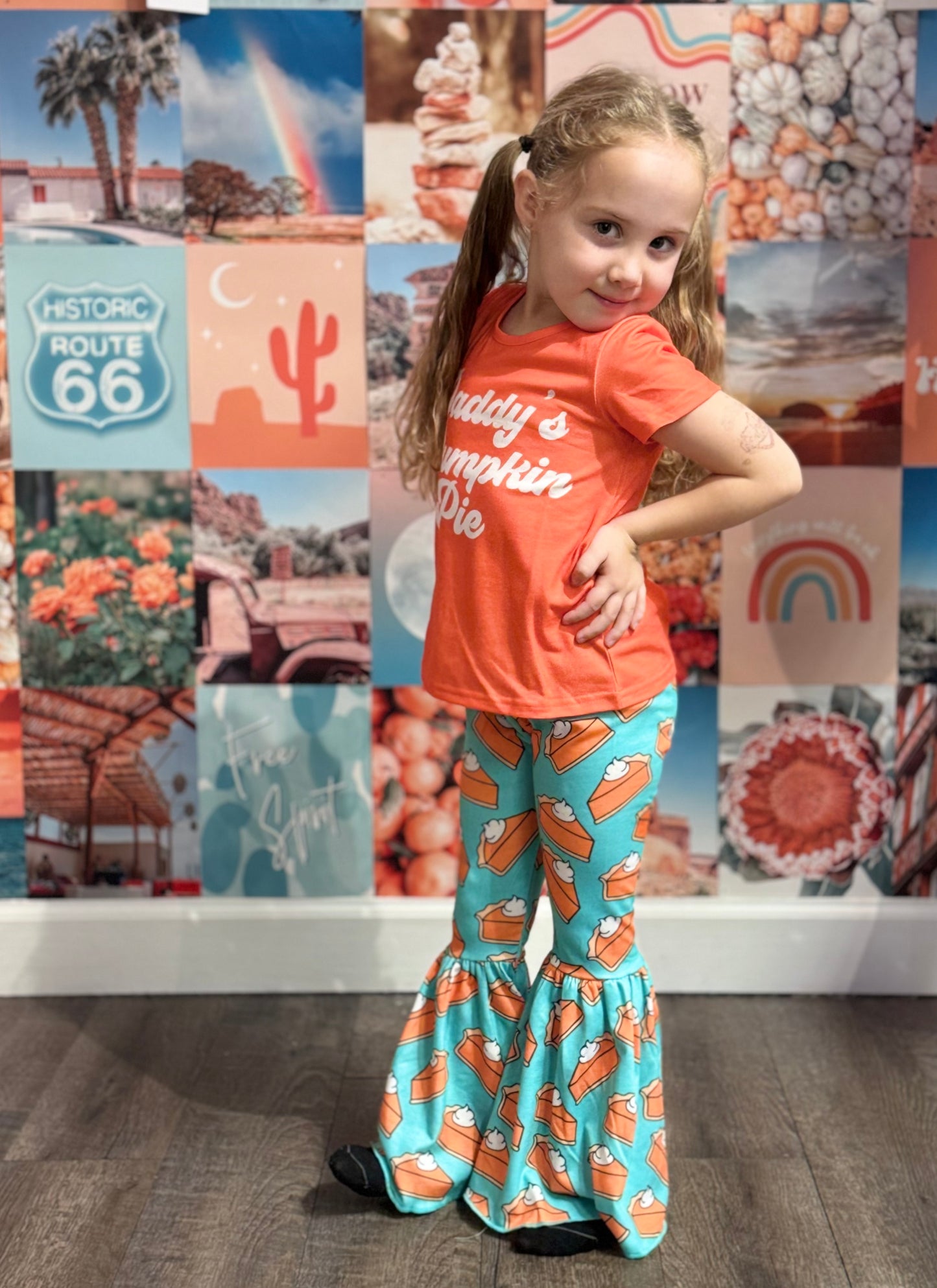 Adorable young girl posing in a festive outfit featuring an orange T-shirt with the text 'Daddy’s Pumpkin Pie' in playful white font. She pairs it with teal bell-bottom pants adorned with a fun pumpkin pie pattern. The background is a vibrant collage of retro-style imagery, including Route 66 signs, rainbows, cacti, and autumn-themed designs.