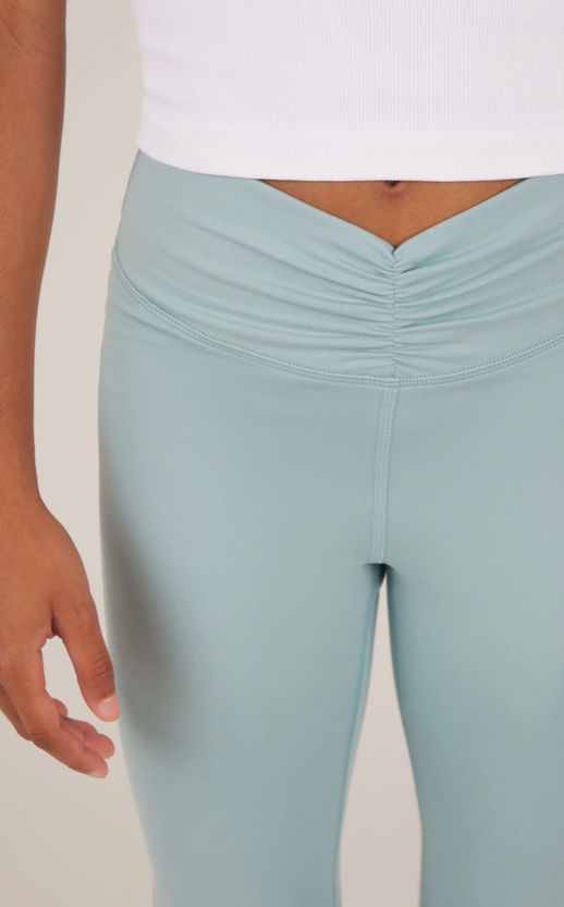 A young model wearing a fitted white short-sleeve crop top paired with high-waisted, flared hot pink yoga pants. The pants feature a ruched crossover waistband for added style and comfort. The outfit creates a sleek and vibrant look, perfect for yoga, dance, or casual athleisure. The model is barefoot, standing in a relaxed pose, with a cheerful expression and natural curly hair styled in a low puff. The plain gray background highlights the outfit's bold colors and clean design.