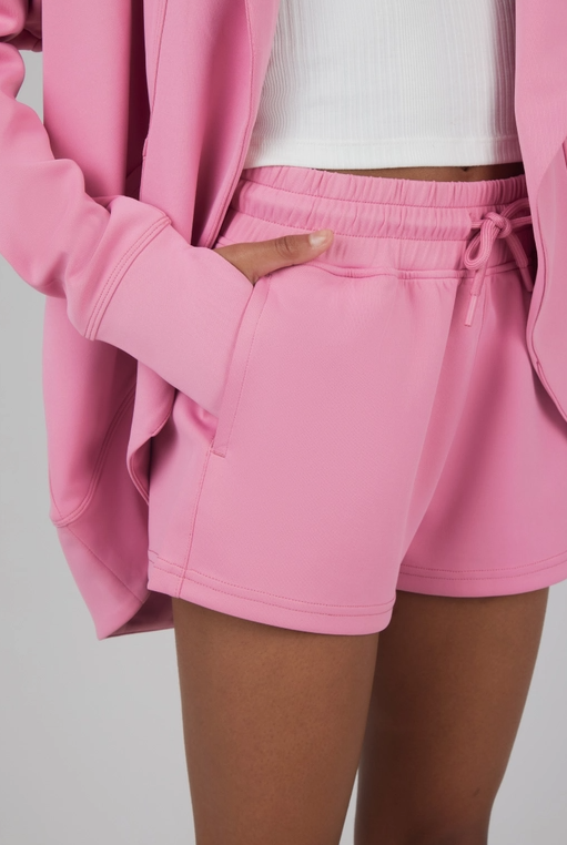 A stylish young girl modeling a casual yet chic outfit consisting of a pink open-front cardigan, matching pink drawstring shorts with pockets, and a white ribbed tank top. The relaxed, coordinated look exudes comfort and versatility, perfect for lounging or casual outings. The soft, pastel tones enhance the outfit's modern and youthful vibe.