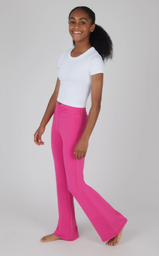 A young model wearing a fitted white short-sleeve crop top paired with high-waisted, flared hot pink yoga pants. The pants feature a ruched crossover waistband for added style and comfort. The outfit creates a sleek and vibrant look, perfect for yoga, dance, or casual athleisure. The model is barefoot, standing in a relaxed pose, with a cheerful expression and natural curly hair styled in a low puff. The plain gray background highlights the outfit's bold colors and clean design.