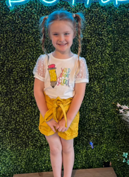 A young girl with braided pigtails smiles brightly while wearing a playful outfit featuring a white top with puffed sheer polka dot sleeves and the text "Wild About School" with a pencil graphic. She pairs the top with mustard-yellow high-waisted shorts tied with a bow. The background showcases lush green foliage with a neon sign, adding a fun, lively atmosphere.
