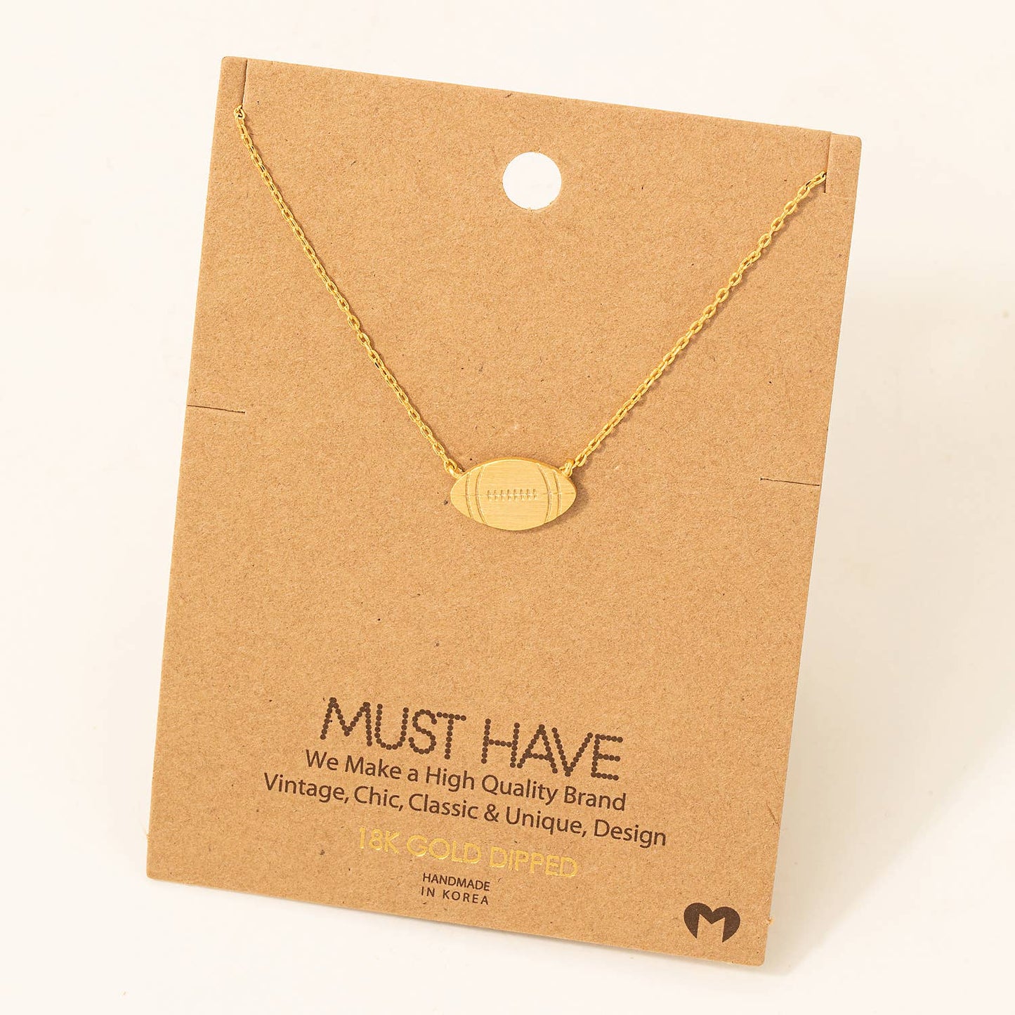 Delicate 18K gold-dipped necklace featuring a minimalist football-shaped pendant with stitched detailing, displayed on a brown cardboard backing with "Must Have" branding. A stylish and chic accessory, perfect for football enthusiasts and game-day fashion.
