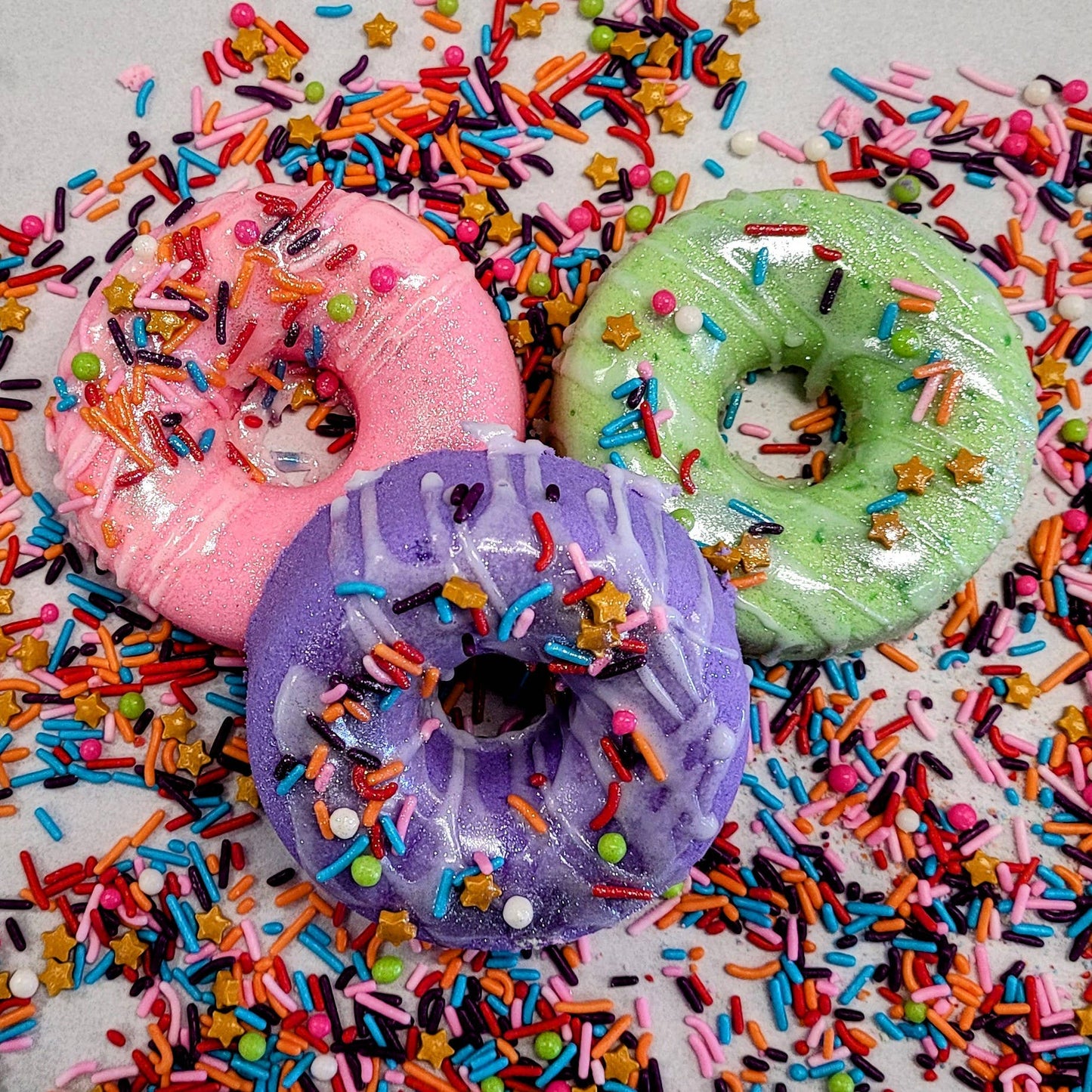 A colorful trio of donut-shaped bath bombs designed to resemble real donuts. Each bath bomb is coated in a shimmery glaze and topped with decorative, brightly colored sprinkles, including stars and round sugar-like beads. The bath bombs come in pastel shades of pink, green, and purple, arranged on a surface scattered with additional sprinkles for a playful and vibrant aesthetic. These whimsical bath bombs combine a fun, dessert-inspired design with a luxurious bath experience.