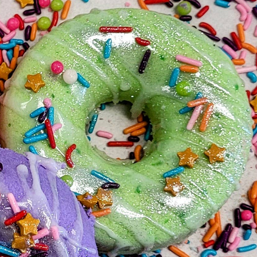 A close-up of a donut-shaped bath bomb in vibrant green, coated with a sparkling shimmer. The bath bomb is decorated with colorful, candy-like sprinkles, including rainbow jimmies, round beads, and small gold stars, giving it a playful, dessert-inspired look. The texture and detailing make it appear almost edible, creating a whimsical aesthetic for a luxurious bath experience.