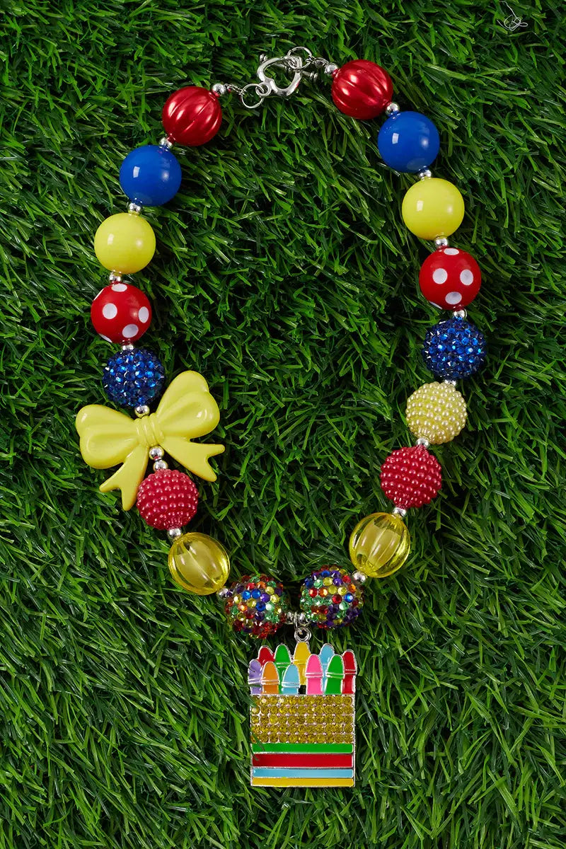 Colorful chunky bead necklace designed for kids, featuring large red, yellow, and blue beads, glittery rhinestone-encrusted beads, and polka-dotted red beads. The necklace includes a bright yellow bow charm and a playful pendant shaped like a box of crayons adorned with multicolored rhinestones. Displayed on a vibrant green grass background, this fun and cheerful accessory is perfect for adding a pop of color to children's outfits.