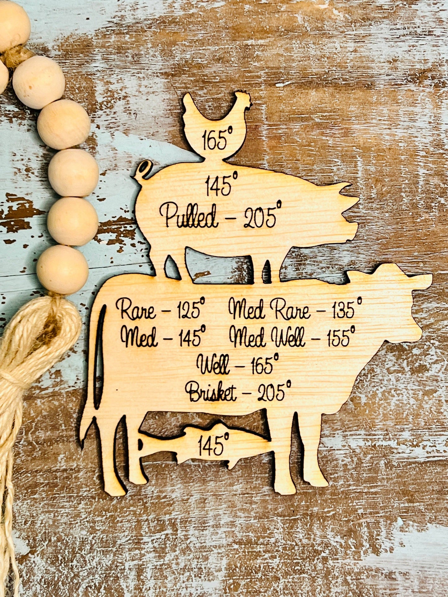 A rustic wooden meat temperature guide featuring engraved silhouettes of a chicken, pig, cow, and fish stacked in a whimsical arrangement. Each animal displays recommended internal cooking temperatures in black script. The chicken notes "165°," the pig lists "145°" and "Pulled – 205°," the cow details a range from "Rare – 125°" to "Brisket – 205°," and the fish is labeled "145°." The background is a weathered, distressed wood surface in shades of blue and brown, with a decorative strand of wooden beads tied