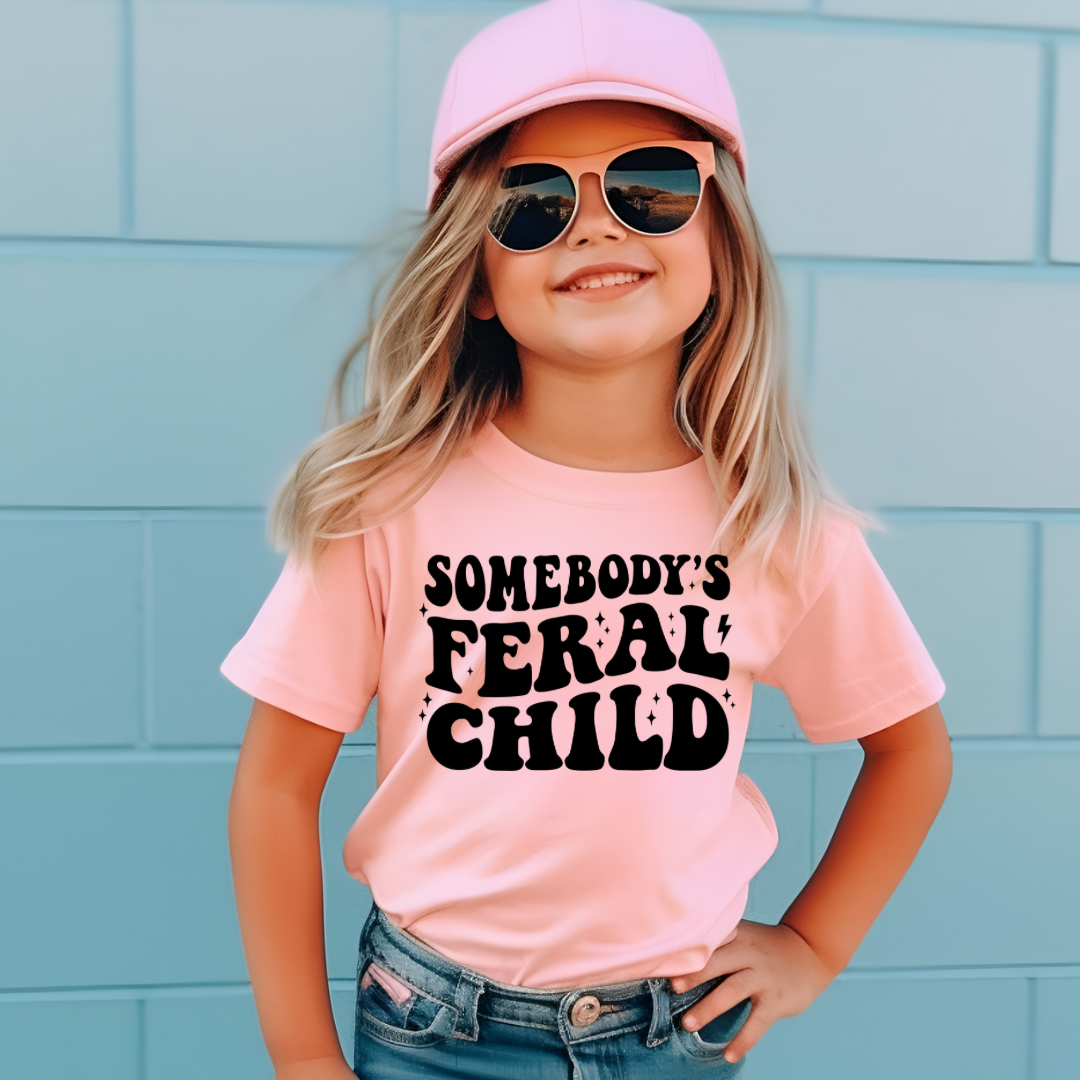 A young girl wearing a light pink t-shirt with bold black text reading "Somebody's Feral Child," paired with denim shorts, a pink cap, and sunglasses. She poses confidently against a light blue wall with a cheerful expression.
