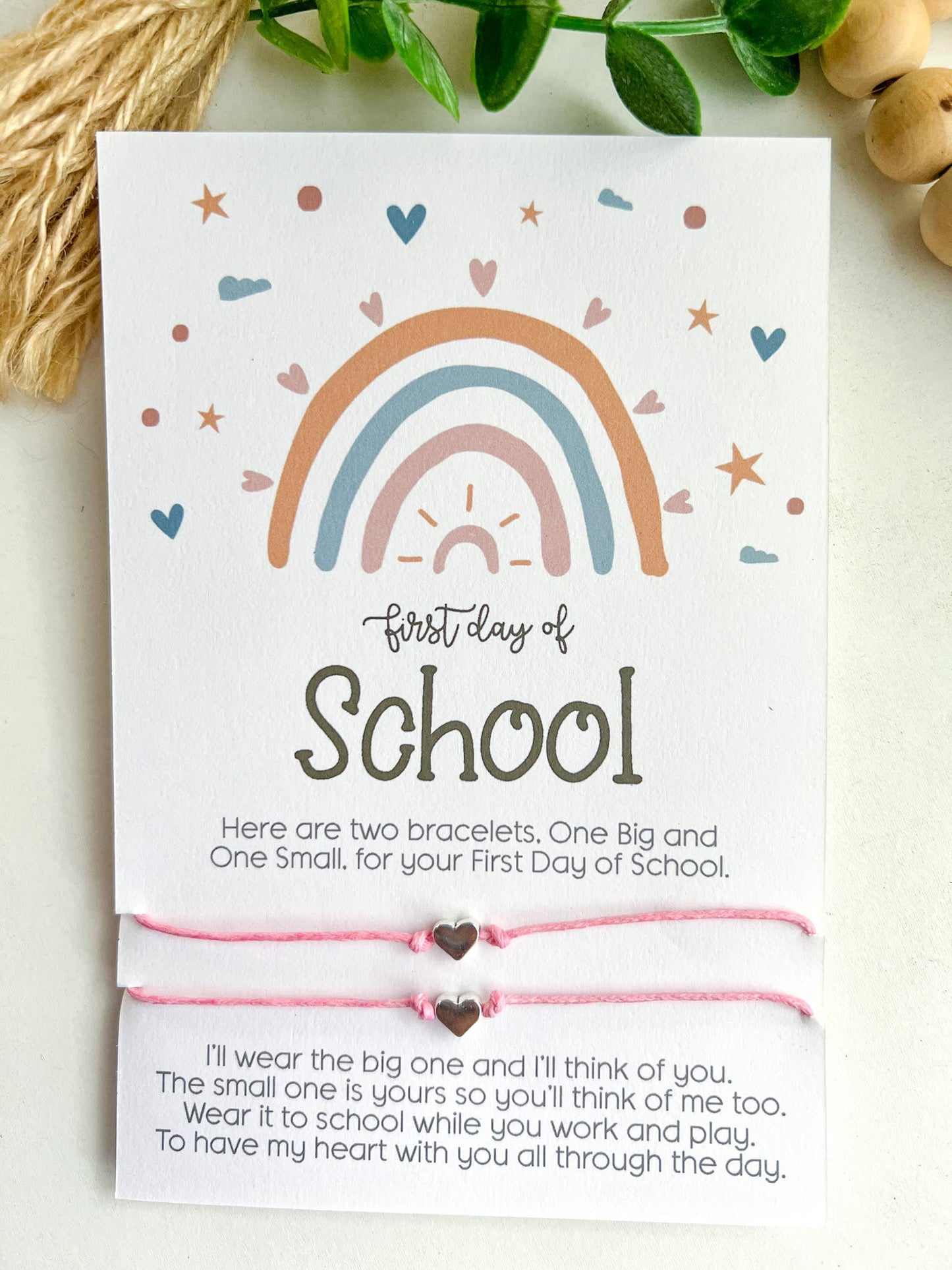 Two "First Day of School" cards featuring a pastel rainbow design with hearts, stars, and clouds. Each card includes a pair of matching string bracelets—one larger and one smaller—with heart-shaped charms in the center. The cards read: "Here are two bracelets. One Big and One Small, for your First Day of School." Below, a heartfelt message explains the bracelets' purpose for connection during the day. One card has mint green bracelets, and the other has pink. Both are placed on a white surface.