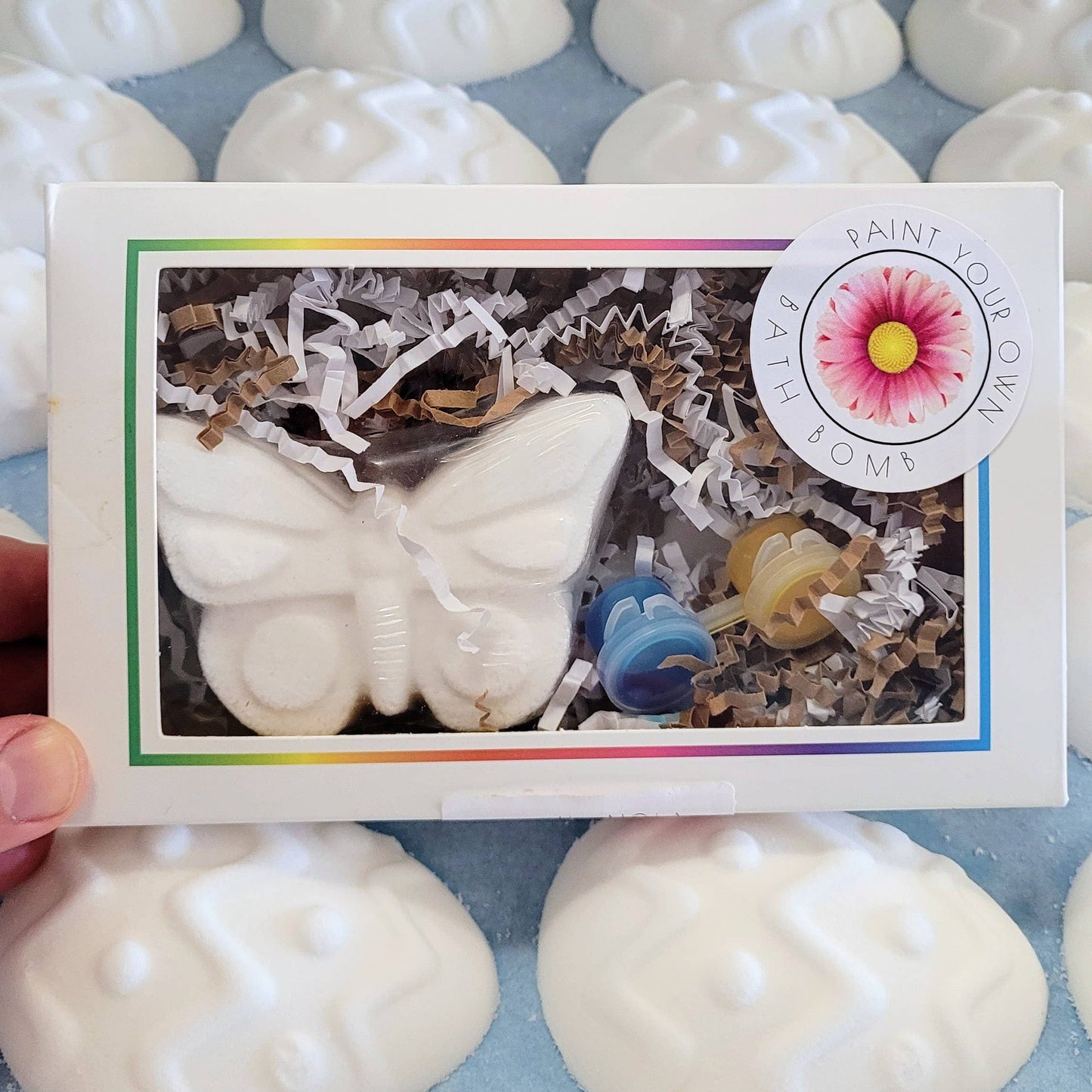 A "Paint Your Own Bath Bomb" kit featuring a butterfly-shaped white bath bomb packaged in a white box with a clear window. The kit includes three small containers of vibrant paint in blue, yellow, and red, nestled among white and brown crinkle paper. A circular sticker with a pink daisy illustration and the text "Paint Your Own Bath Bomb" adds a creative, DIY-themed touch. The background shows additional plain white bath bombs, emphasizing the customizable nature of the product. Perfect for gifting or craft
