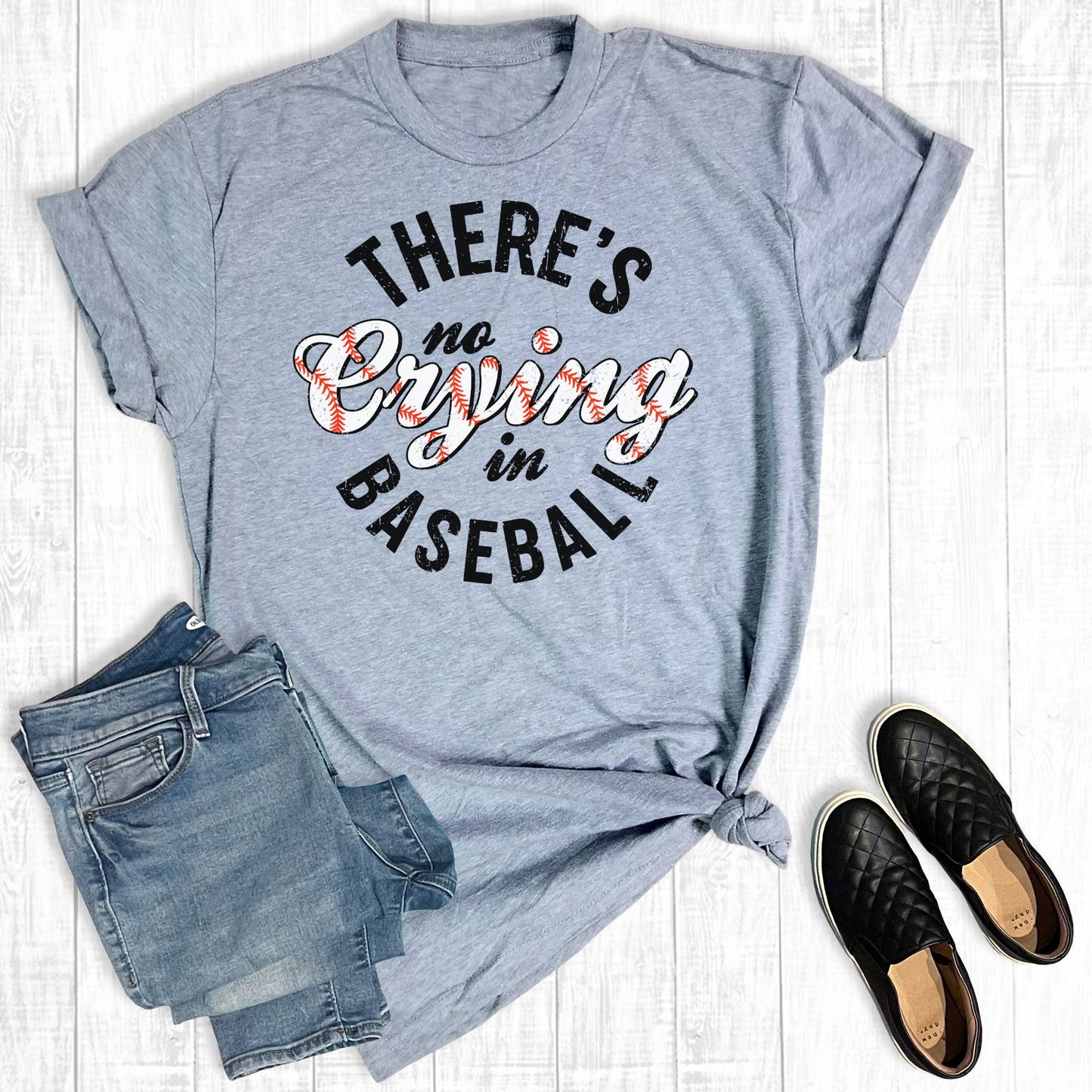 A heather grey t-shirt with the phrase "There's no crying in baseball" printed in black and white with red baseball stitching details. The shirt is styled with a knot on one side, displayed next to a pair of denim shorts and black quilted slip-on shoes on a light wooden surface.
