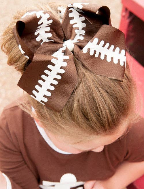 Large brown grosgrain hair bow designed with white football lace detailing, perfect for game-day or sports-themed events. The bow is styled on a decorative background and shown in use atop a child's hairstyle, complementing a sporty outfit. Ideal for football fans and festive occasions.