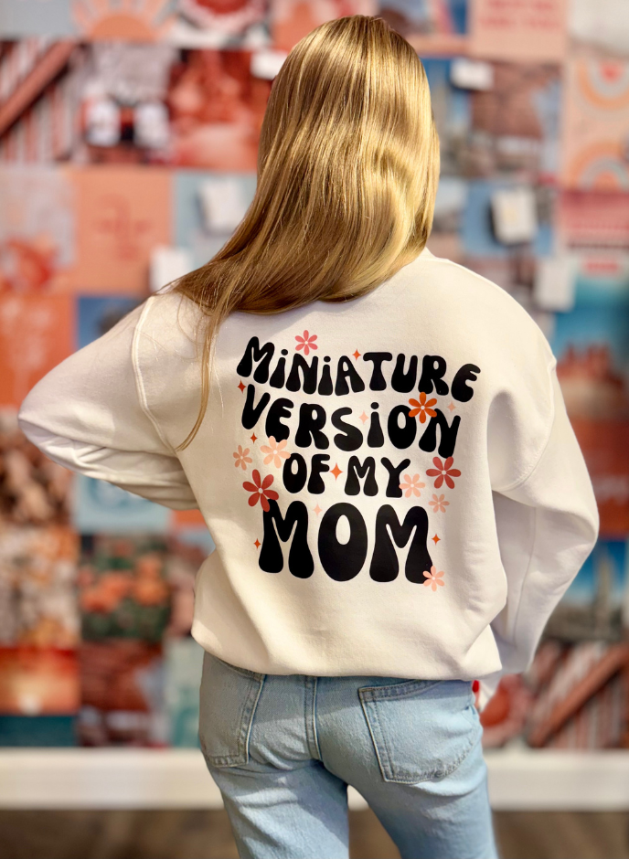 Back view of a white sweatshirt with 'Miniature Version of My Mom' in bold, playful lettering and floral details.