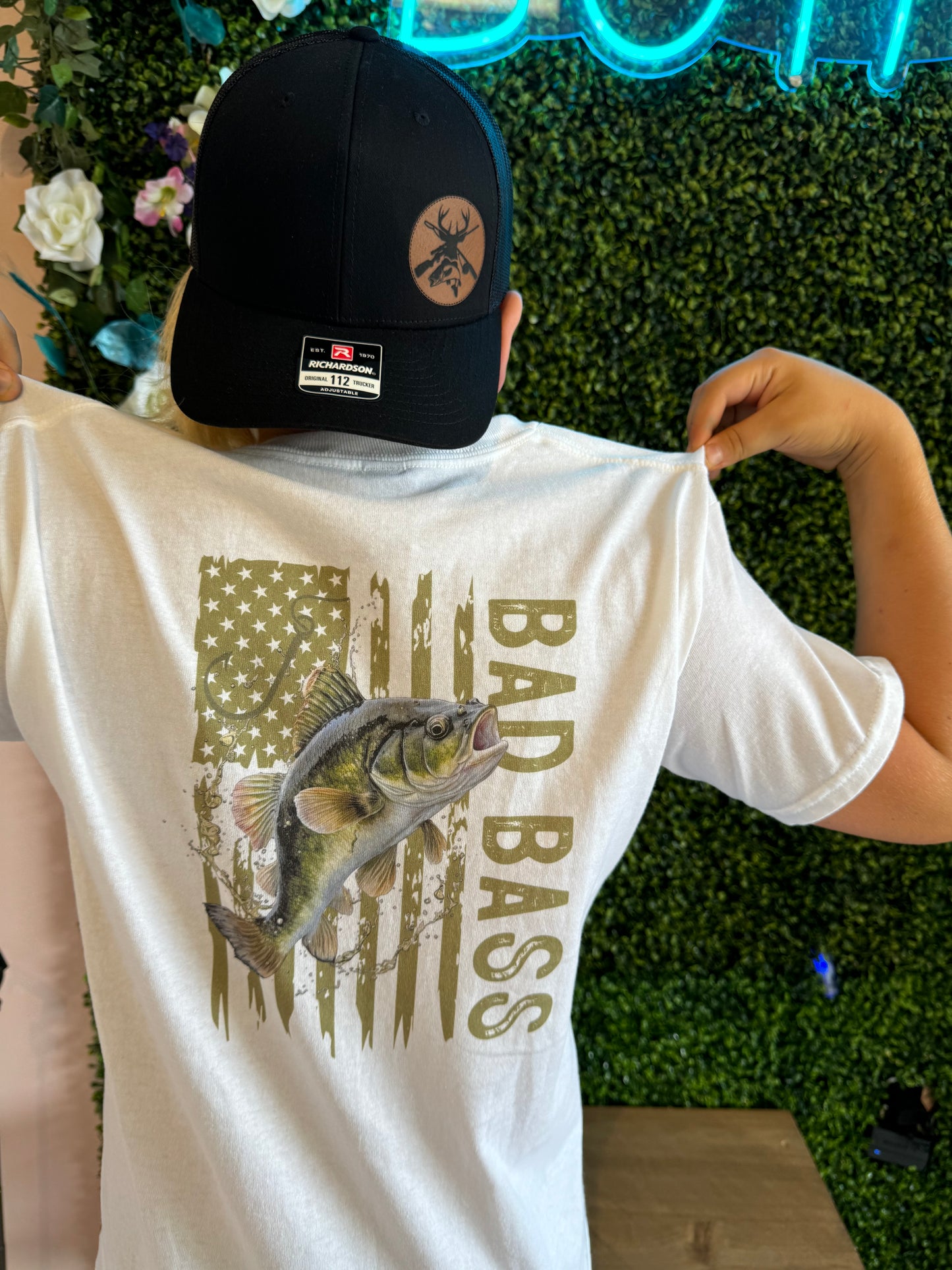 Back view of a person wearing a white T-shirt with a graphic design featuring a large bass fish over a distressed American flag pattern and the text 'Bad Bass' in bold. The individual is also wearing a black baseball cap with a tan patch featuring a deer skull design. The background consists of lush green foliage and neon blue lighting, creating a modern and bold aesthetic.