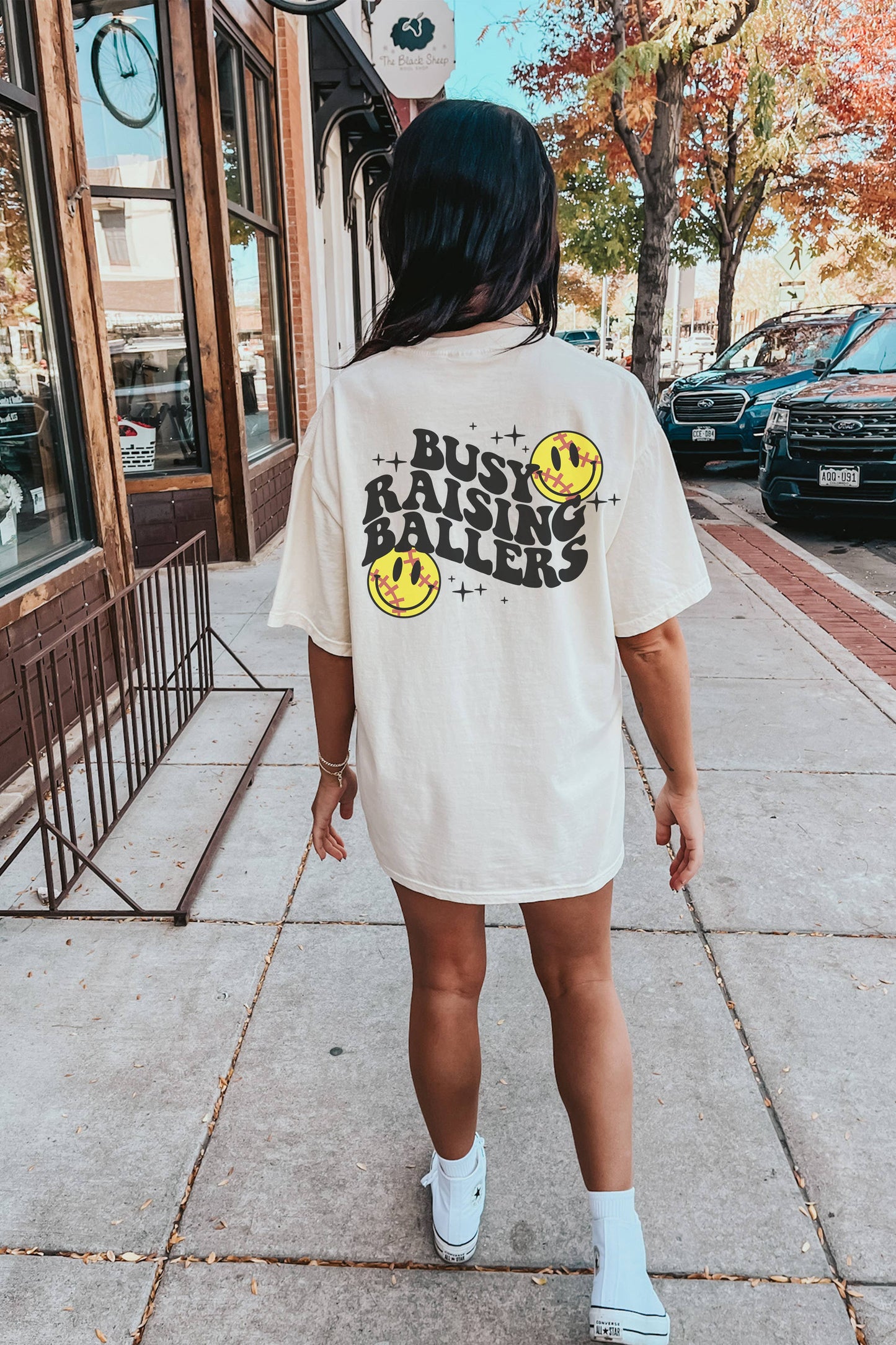 The back of the shirt showcases bold black text reading "BUSY RAISING BALLERS" surrounded by two softball graphics and sparkles. The playful design emphasizes a sporty, family-oriented theme, making it perfect for softball moms or sports enthusiasts. The casual backdrop of a sidewalk with parked cars and fall foliage enhances the relaxed, everyday appeal of the outfit.