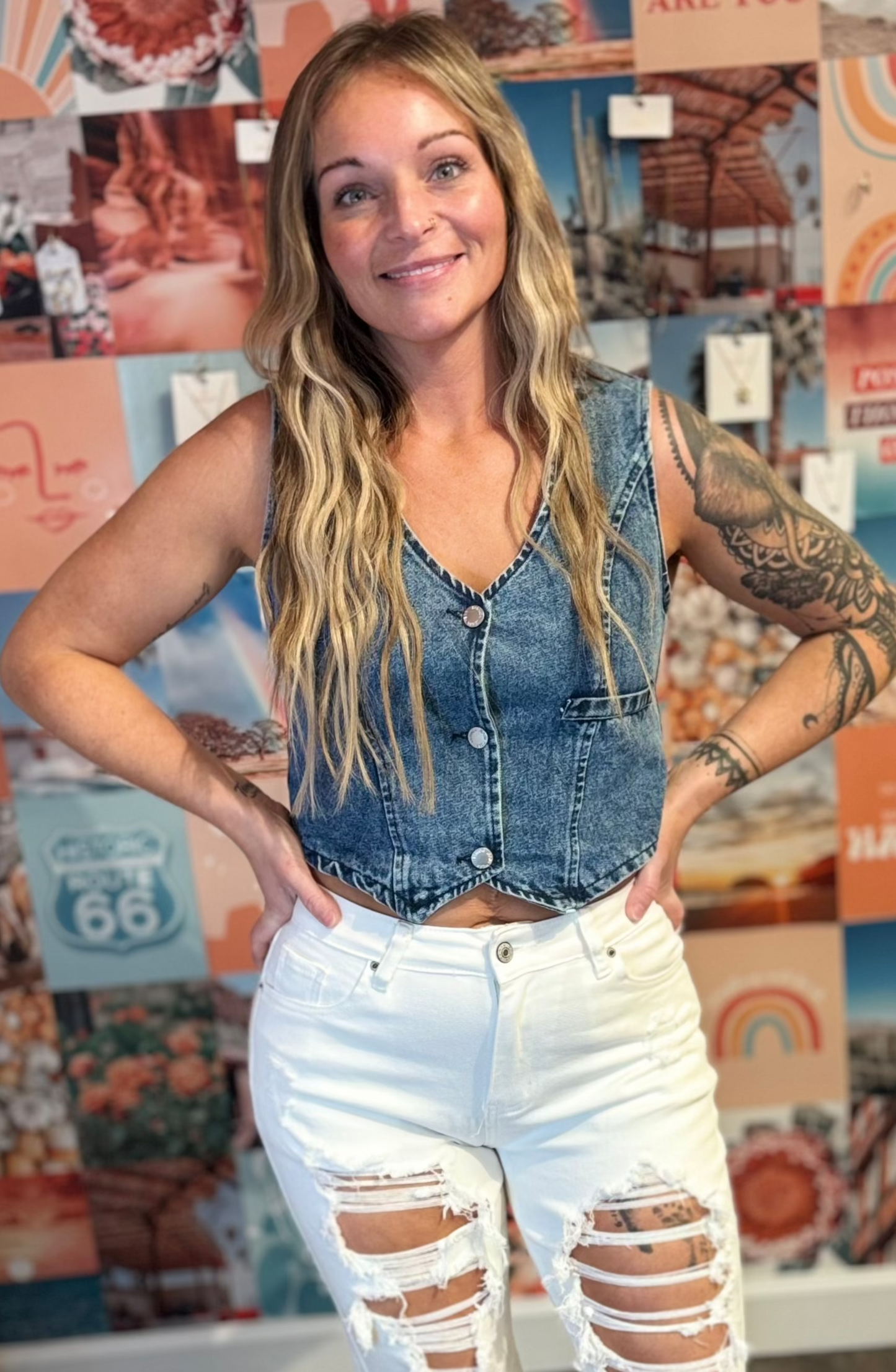 Woman in a sleeveless denim vest and white distressed jeans poses confidently in front of a vibrant, bohemian-inspired wall. Her outfit highlights casual Western-inspired style with modern, trendy elements.