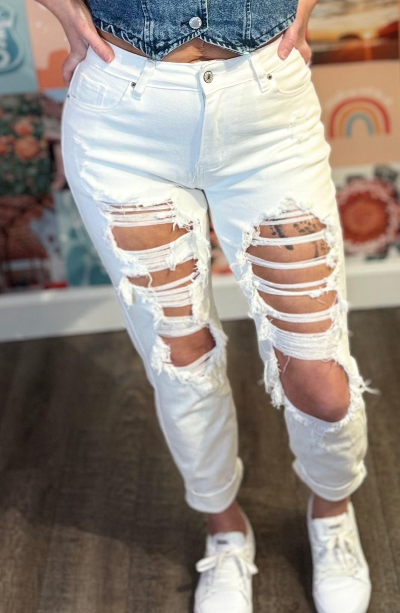 A close-up view of a pair of high-waisted, white distressed boyfriend jeans featuring extensive ripped detailing across the thighs, knees, and lower legs, giving them a bold, edgy look. The jeans have a button and zip closure with front and back pockets, and the hem is cropped and slightly rolled.