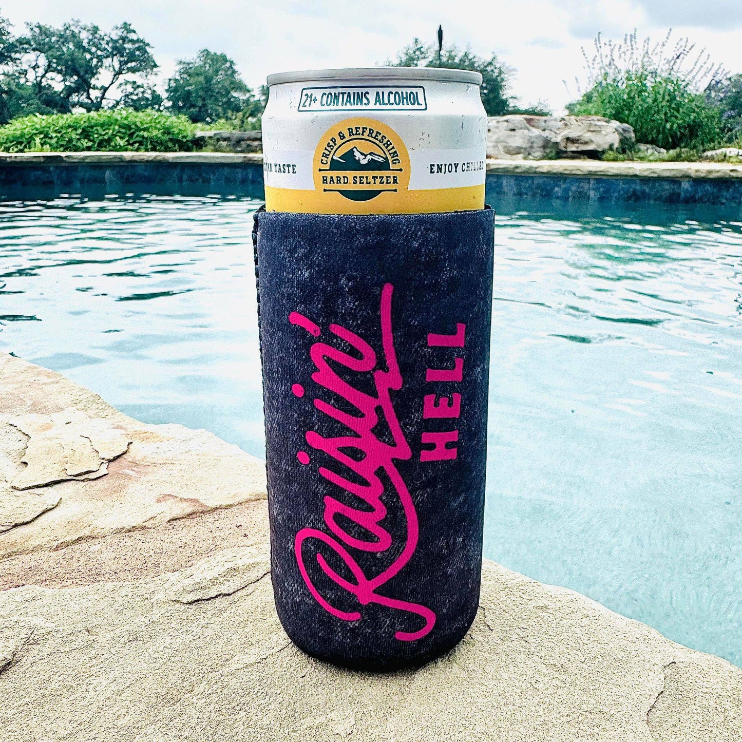 A slim can koozie with the phrase "Raisin' Hell" written in bold pink script on a dark background, holding a can of hard seltzer. The can displays a yellow and white label with text such as "Crisp & Refreshing Hard Seltzer" and "Enjoy Chilled." The koozie is placed on a stone poolside, with a serene background featuring a turquoise pool, lush greenery, and cloudy skies. The scene exudes a laid-back, summer vibe, perfect for relaxing outdoors with a drink.