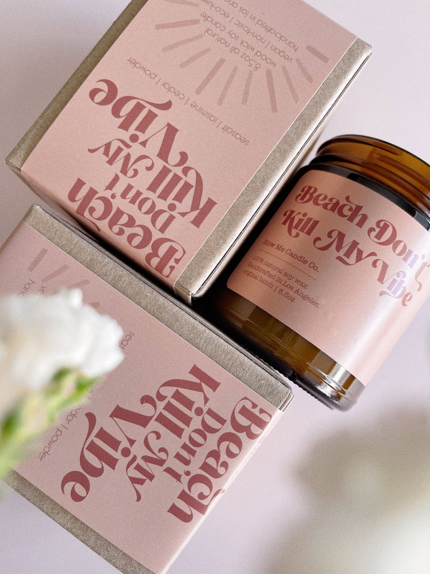 Close-up of a luxury soy candle by Blow Me Candle Co., featuring the label 'Beach Don’t Kill My Vibe' in retro-style pink lettering on a muted pink box. The candle scent includes notes of seasalt, jasmine, cedar, and powder. The glass jar candle with a wooden wick is elegantly packaged, perfect for gifting or home decor.