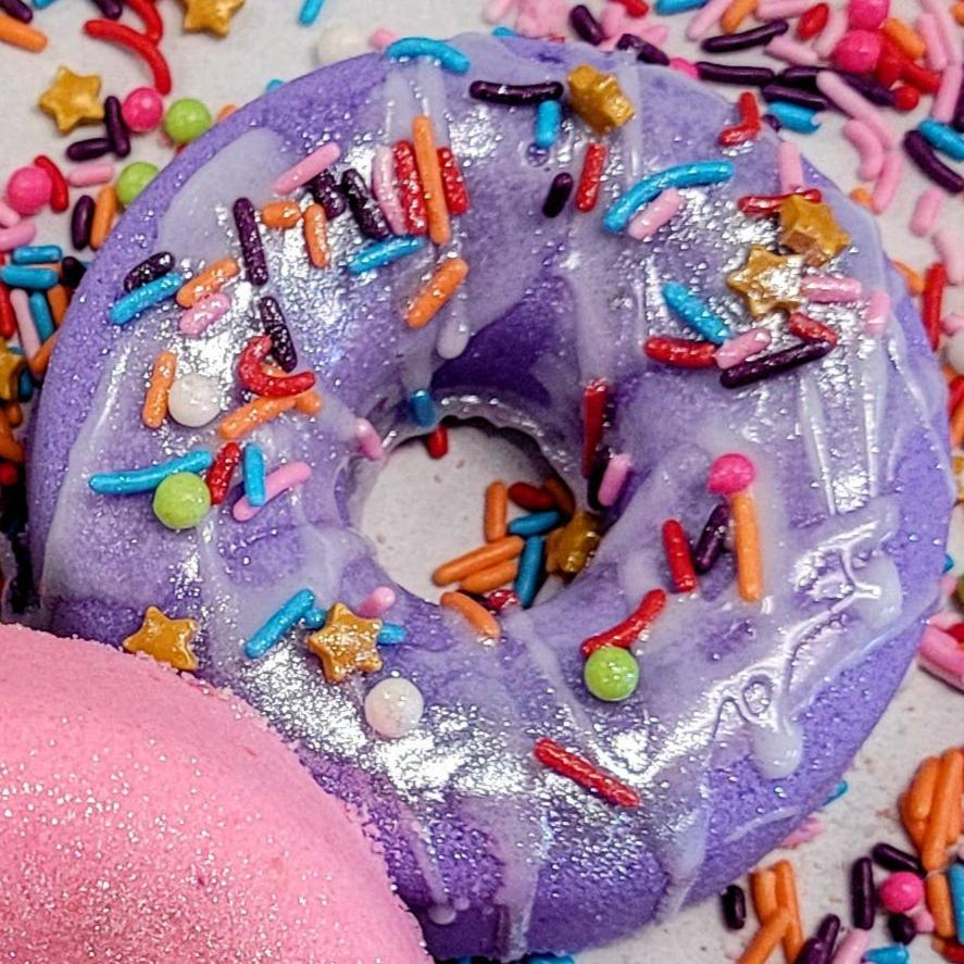 A close-up of a donut-shaped bath bomb in vibrant purple, coated with a sparkling shimmer. The bath bomb is decorated with colorful, candy-like sprinkles, including rainbow jimmies, round beads, and small gold stars, giving it a playful, dessert-inspired look. The texture and detailing make it appear almost edible, creating a whimsical aesthetic for a luxurious bath experience.