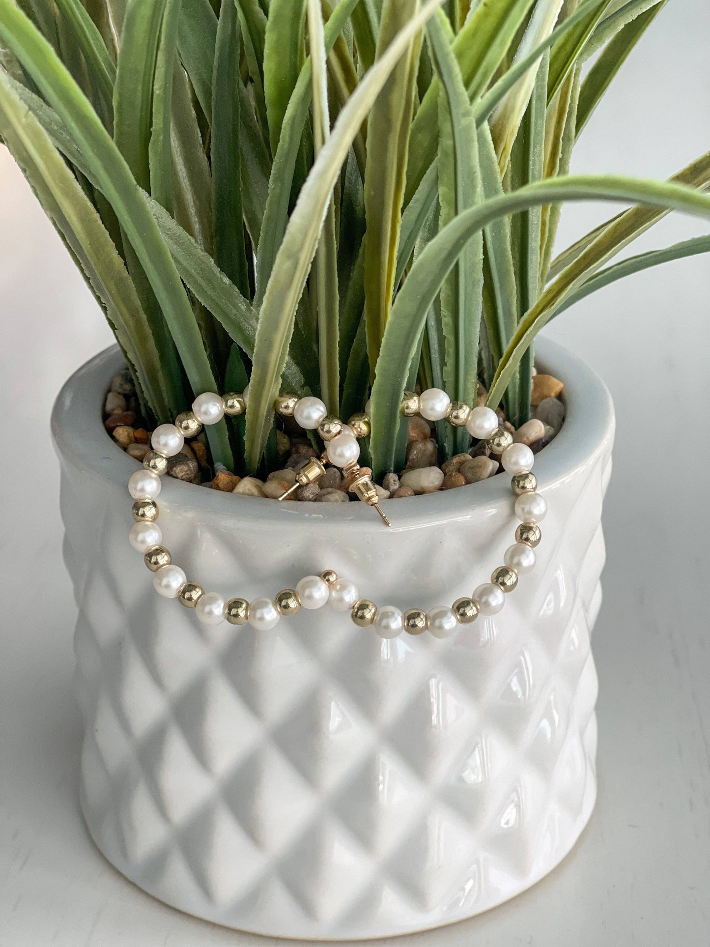 A pair of stylish hoop earrings featuring a mix of glossy white faux pearls and shiny gold beads, arranged in an alternating pattern. The earrings are displayed on a white surface with a decorative sprig of orange and yellow faux berries in the background, adding a warm, autumnal touch to the composition.
