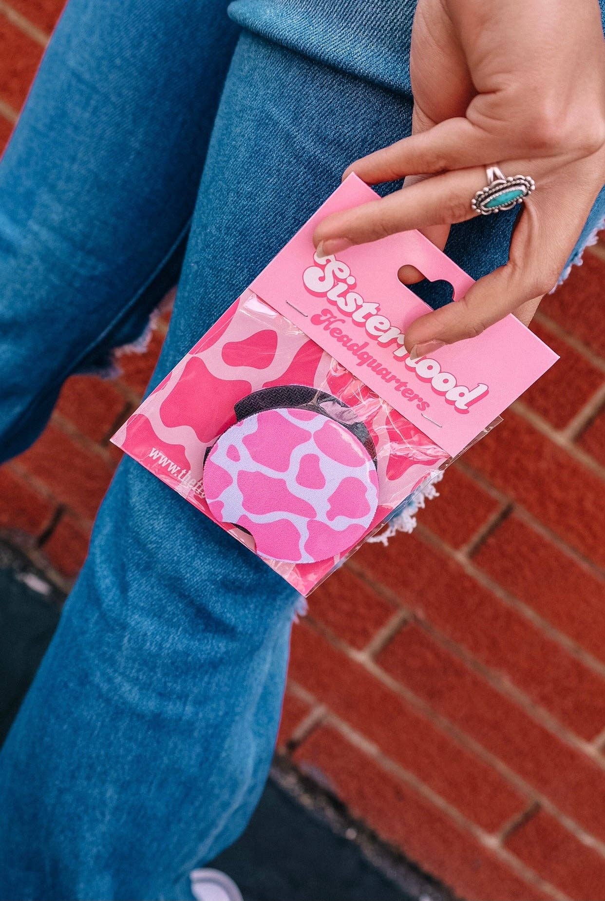 "Close-up of a hand holding a packaged set of pink cow-print cup holder coasters labeled 'Sisterhood Headquarters,' with a backdrop of distressed denim and a brick wall."