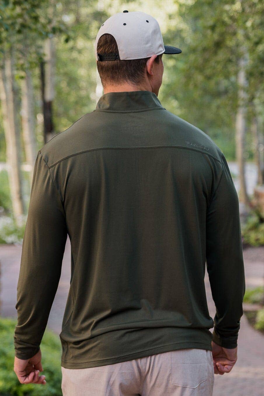 Rear view of a man in a burlebo olive-green long-sleeve quarter-zip pullover shirt, paired with a light gray and black logo cap, standing in a natural wooded area.