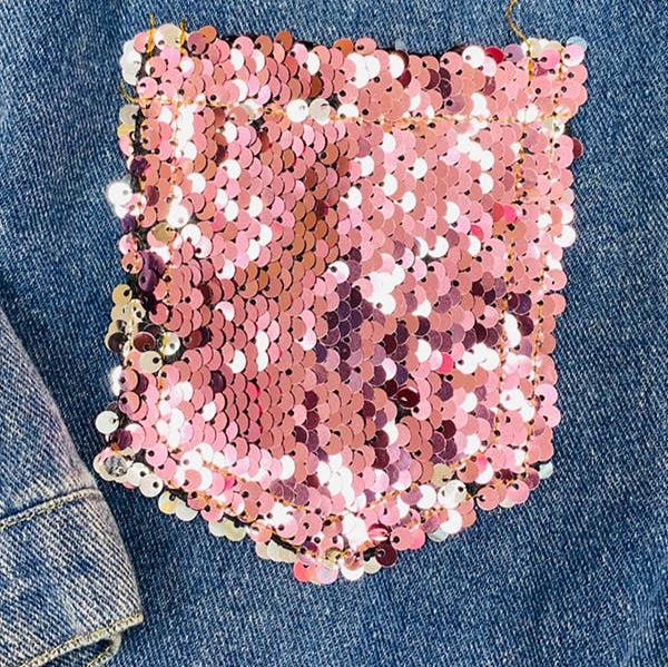 Trendy girls' denim jacket featuring pink sequin embellishments on the front pockets and distressed detailing for a stylish, modern look. Paired with brown suede fringe boots with gold stud accents, this outfit is perfect for casual outings, fall fashion, or statement kid’s clothing. Set against a rustic wood backdrop for a chic presentation.
