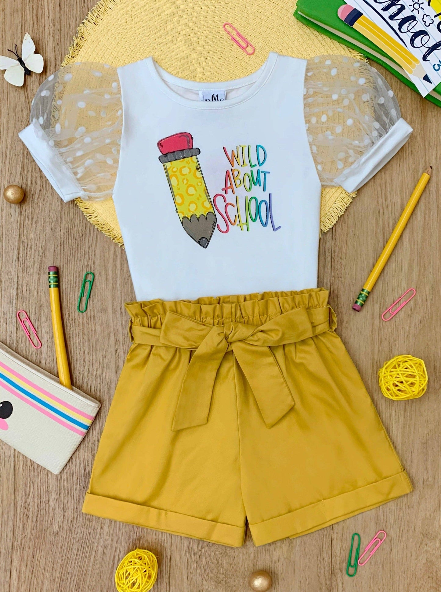 A vibrant children's outfit featuring a white top with puffed sheer polka dot sleeves and colorful text reading "Wild About School" alongside a playful pencil graphic. Paired with mustard-yellow high-waisted shorts tied with a bow. The ensemble is styled with school-themed props like pencils, paper clips, and a playful pouch, set on a wooden surface.