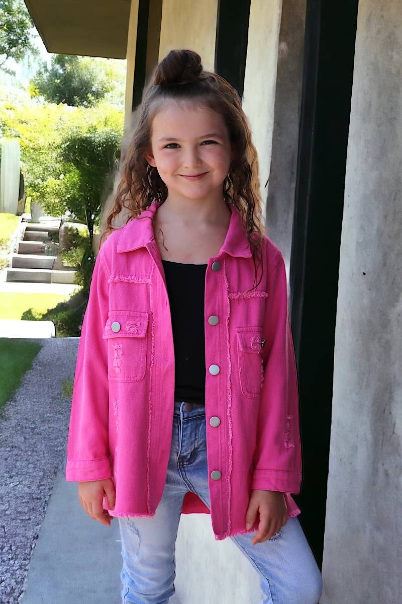Smiling young girl wearing a bright pink distressed shacket over a black top, paired with light-wash denim jeans. Styled outdoors with a modern architectural backdrop, this outfit is a perfect mix of bold and casual fashion for kids.