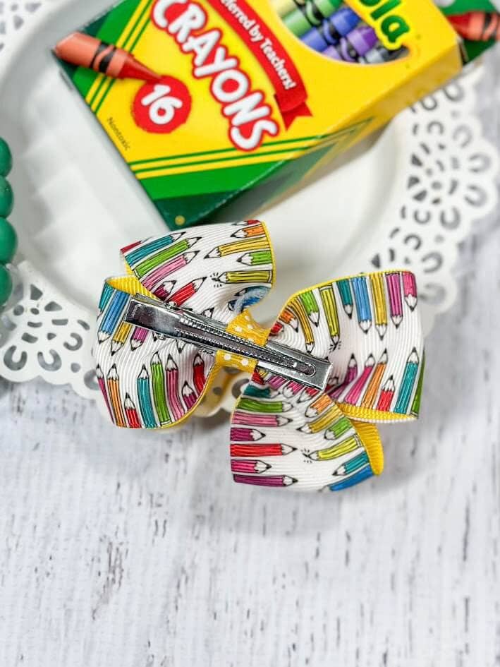 Alt text: A vibrant school-themed hair bow featuring a yellow ribbon layered with a patterned ribbon displaying colorful pencils. The bow is adorned with an enamel charm of stacked books labeled "Art," "Math," and "Reading," topped with a red apple. The bow is displayed on a decorative white plate, accompanied by a green beaded bracelet and a box of Crayola crayons in the background, adding to the back-to-school theme.