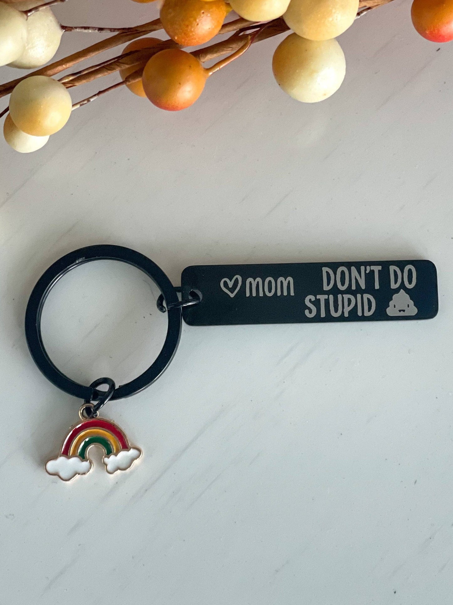 Black keychain with engraved text reading "♥ mom DON’T DO STUPID" alongside a small illustration of a car. Attached is a colorful enamel charm featuring a rainbow with clouds. A playful, practical accessory with a heartfelt and humorous reminder.