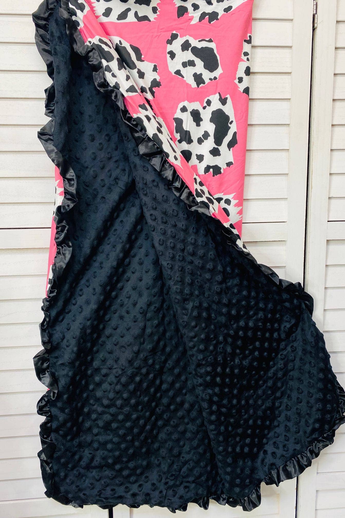 "Pink and white cow-print blanket with a luxurious black minky interior and satin ruffle trim, displayed against a white wooden backdrop."