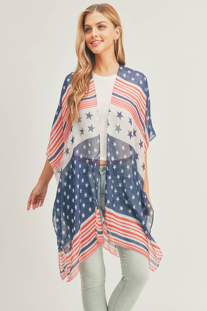 A woman wearing a lightweight patriotic shawl with a design featuring blue with white stars and red and white stripes. The shawl drapes elegantly over her shoulders, complementing her white ribbed top. She has long, wavy blonde hair and is accessorized with gold hoop earrings, posing against a neutral background.
