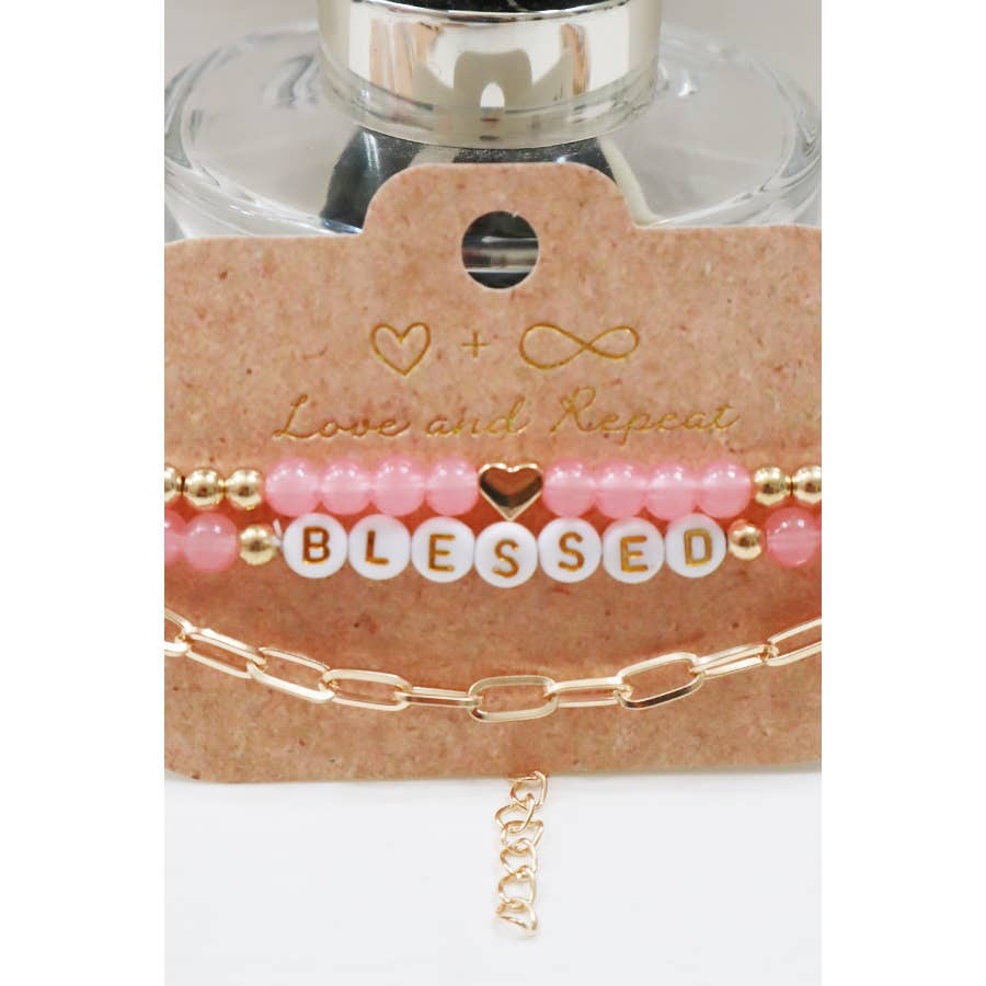 Set of elegant bracelets featuring gold beads, pink beads, and a white letter bead design spelling 'BLESSED.' The set includes a dainty gold heart accent and a stylish gold chain bracelet, displayed on a rustic beige backing card with a minimalist heart design, perfect for gifting or accessorizing with a positive touch.