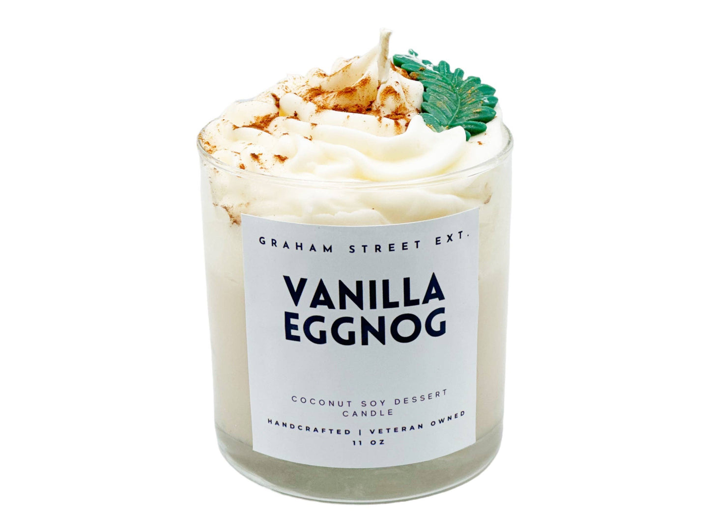 A decorative candle in a clear glass jar labeled "Graham Street Ext. Vanilla Eggnog." The candle features a whipped topping design, sprinkled with cinnamon and adorned with a green leaf-shaped embellishment, evoking the look of a festive dessert. The label indicates it is a coconut soy dessert candle, handcrafted by a veteran-owned business, with a capacity of 11 oz.